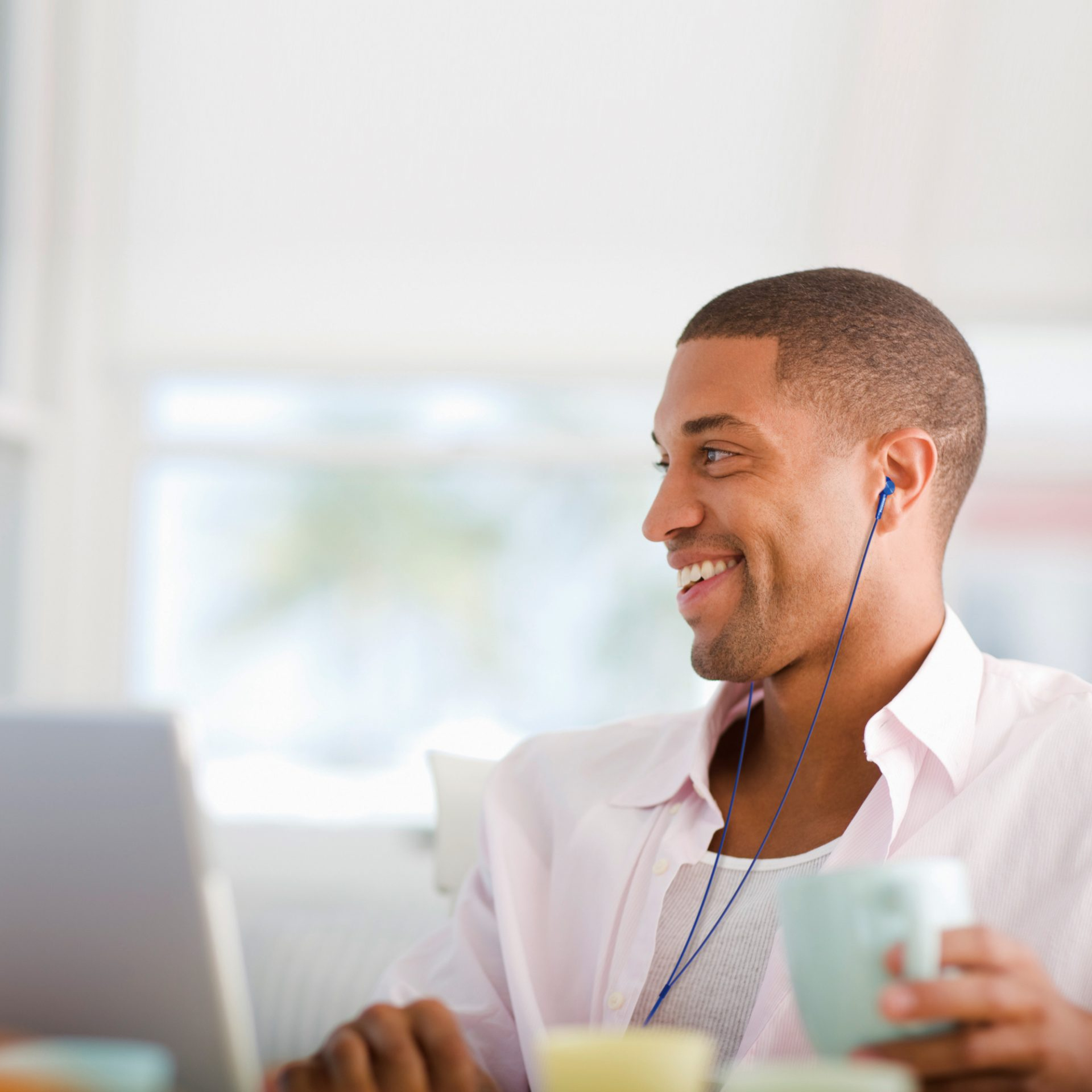 Uśmiechnięty mężczyzna ze słuchawkami trzyma kubek i używa laptopa. Ma na sobie różową koszulę, a tło jest rozmyte.