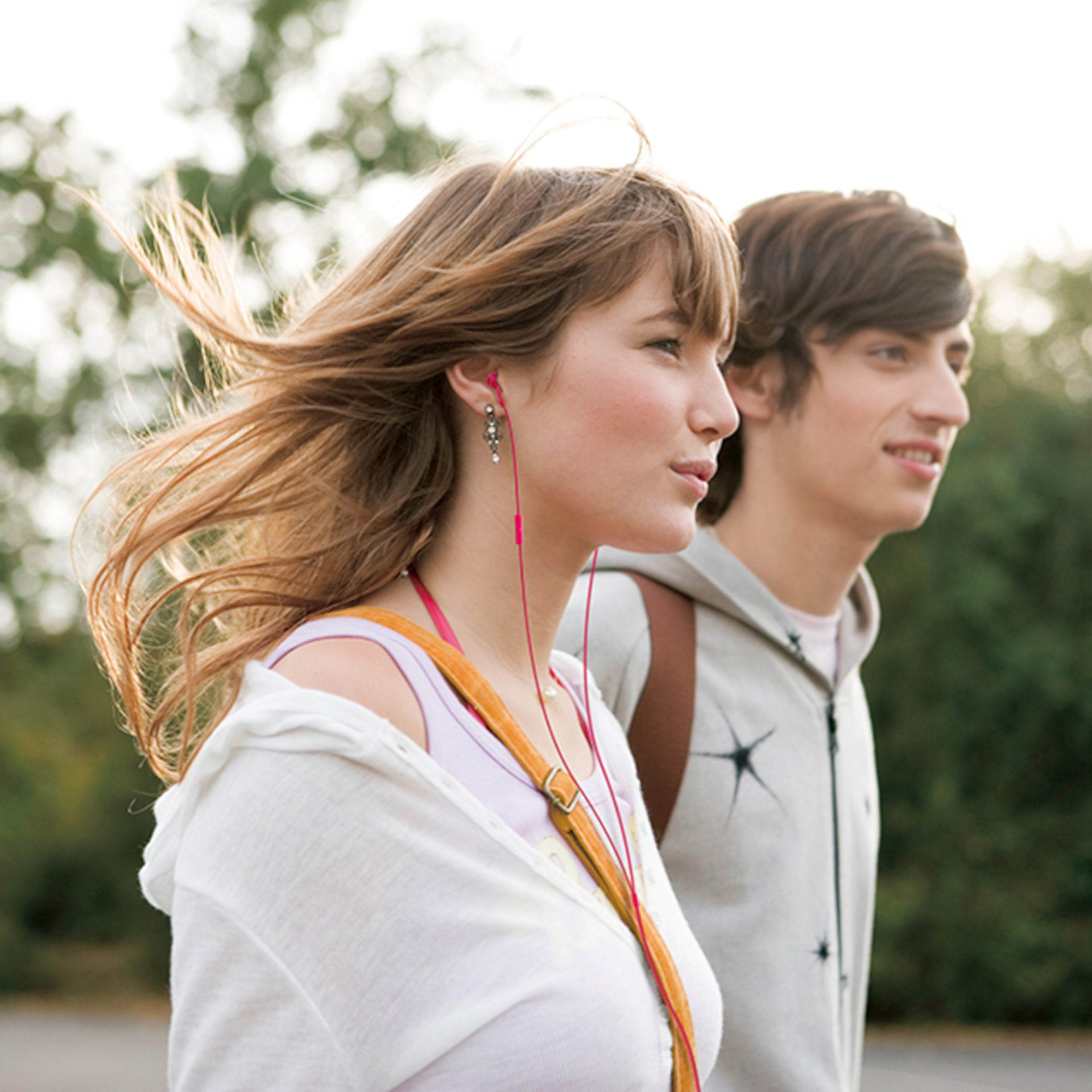 Kobieta z powiewającymi włosami i różowymi słuchawkami idzie z mężczyzną w bluzie z kapturem. W tle zielone drzewa.