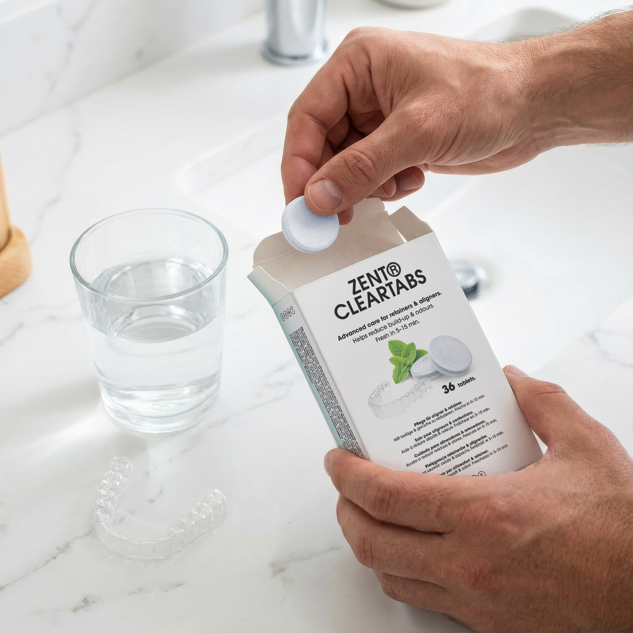 Ręka umieszcza tabletkę z pudełka z napisem ZENT® CLEARTABS. Szklanka wody i aparat retencyjny na powierzchni.