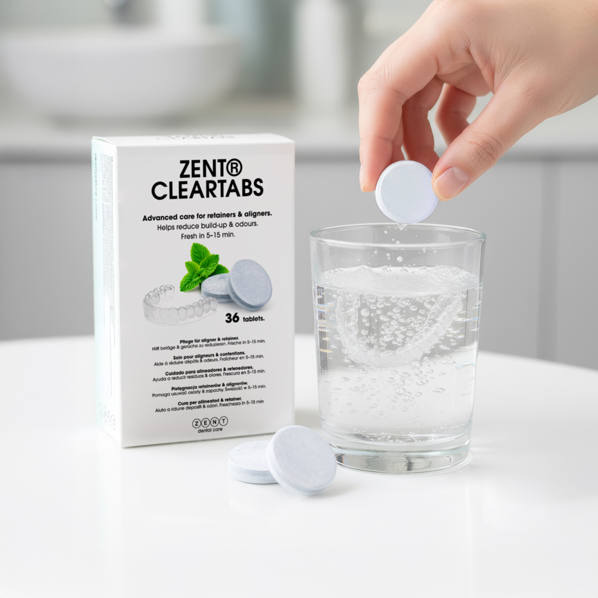 Dłoń umieszcza tabletkę w szklance wody, obok pudełka ZENT Cleartabs i innych tabletek. Woda musuje.