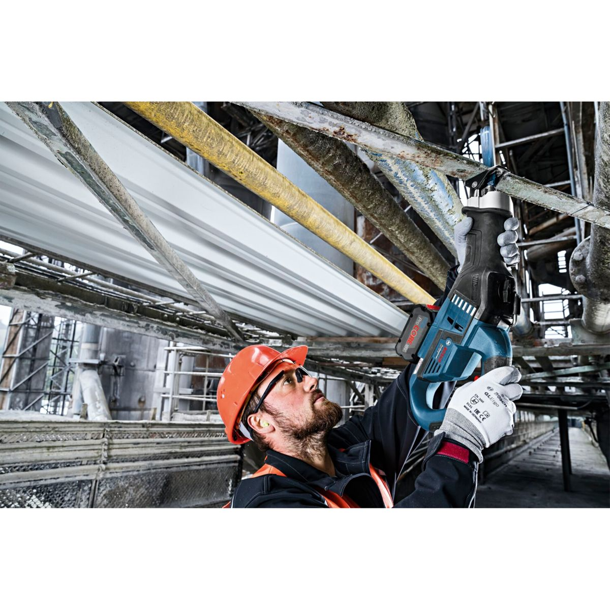Ein Arbeiter mit Schutzhelm verwendet eine Säge an Metallträgern. Industrielle Umgebung mit Rohren und Strukturen im Hintergrund.