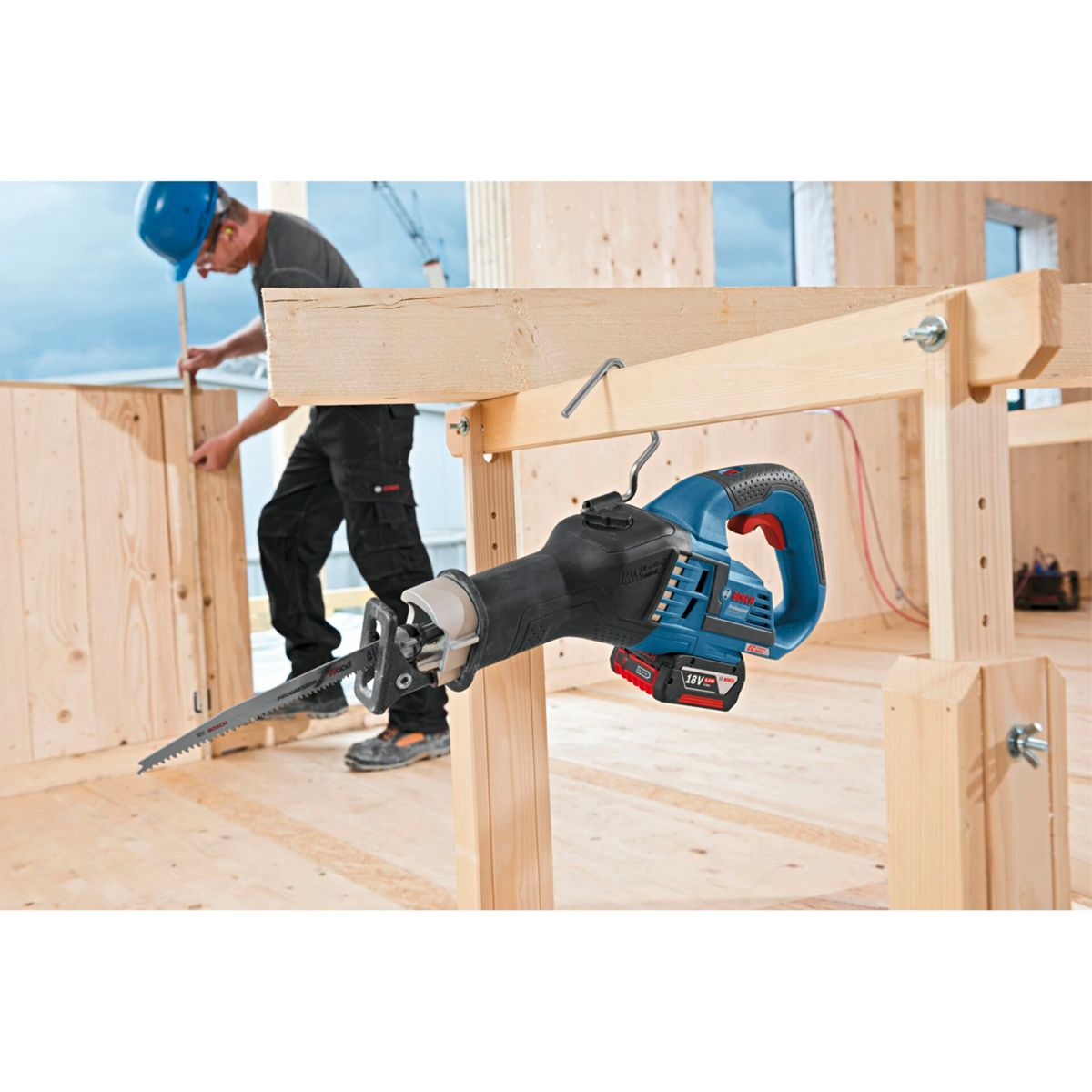 Eine blau-schwarze Säbelsäge hängt an einem Holzrahmen. Ein Bauarbeiter arbeitet im Hintergrund an einer Holzkonstruktion.
