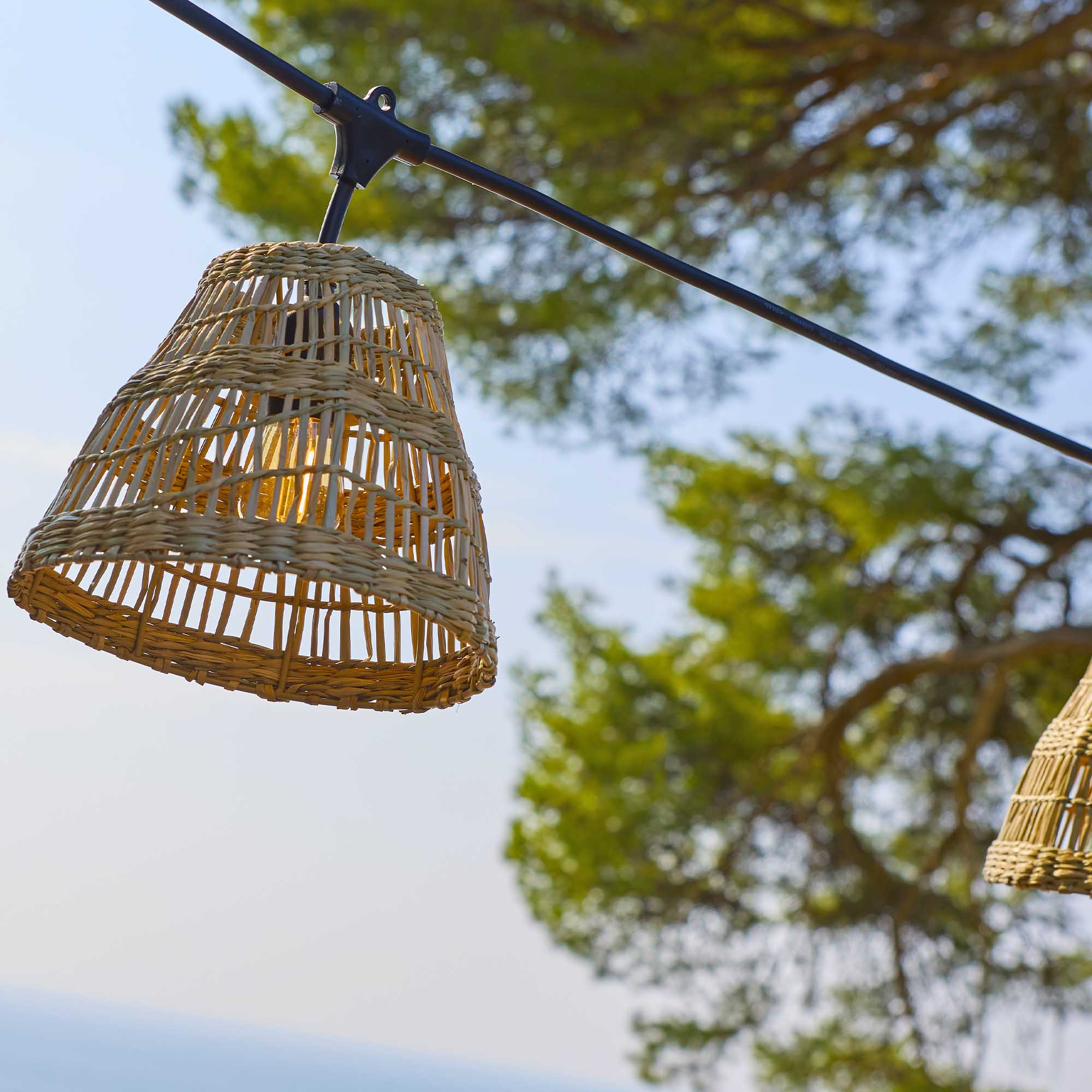 Ein gewebter Lampenschirm hängt an einem Draht, mit einem Baum im Hintergrund. Der Himmel ist blau.