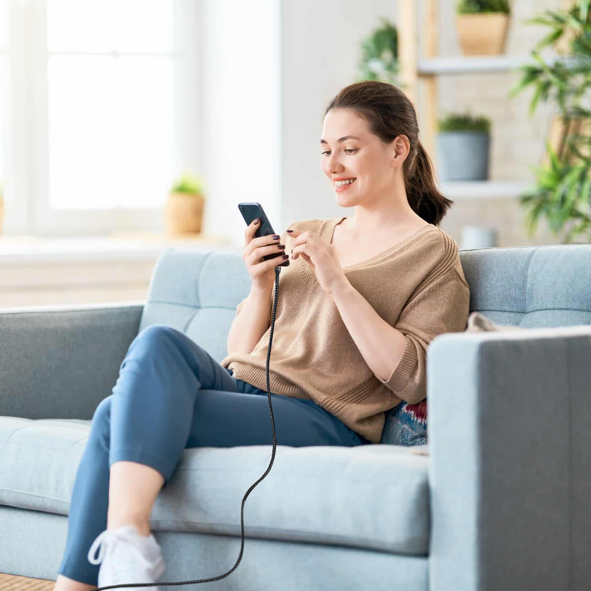 Eine Frau sitzt auf einem blauen Sofa und benutzt ein Smartphone. Sie trägt einen braunen Pullover und blaue Hose. Ein Kabel ist angeschlossen.