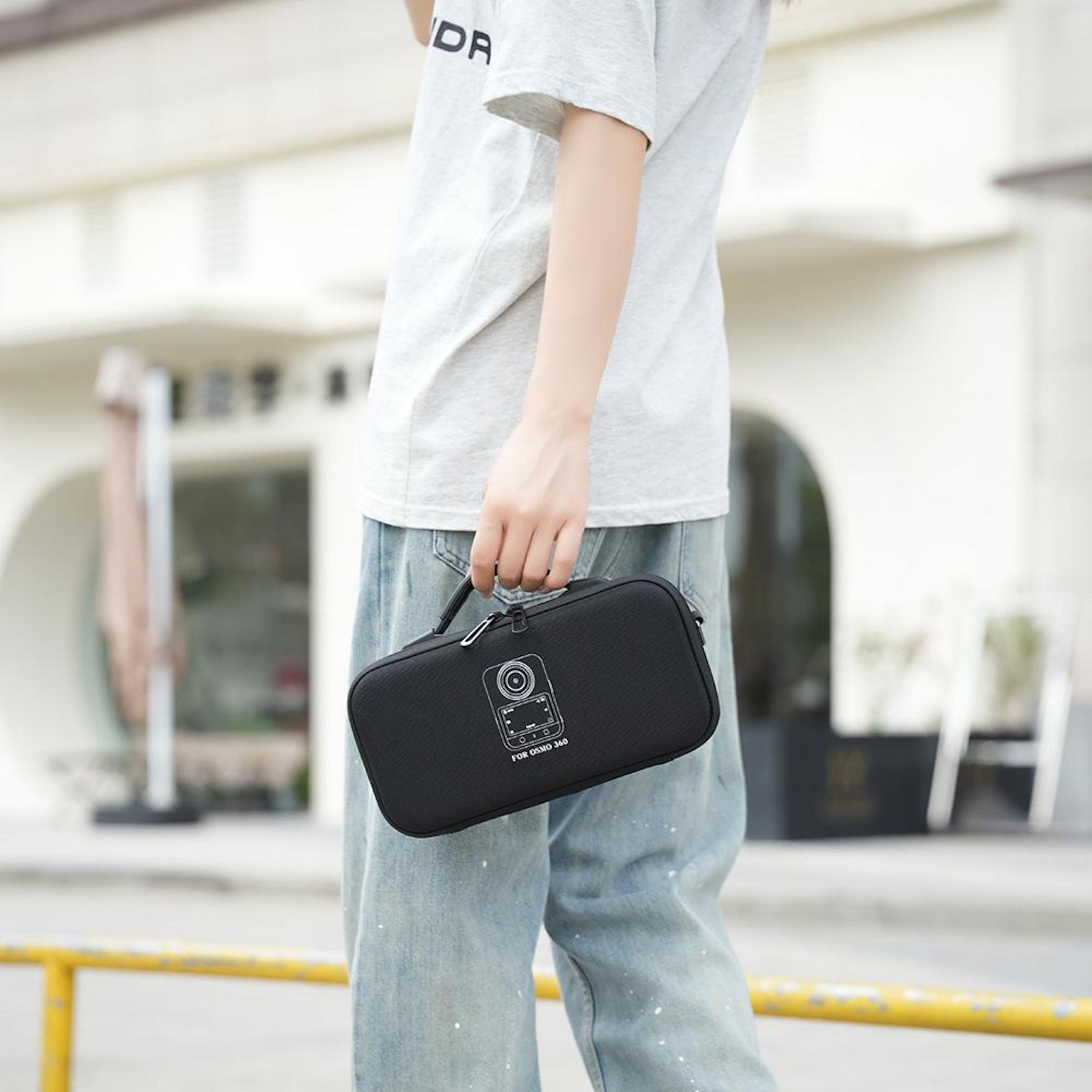 Person hält eine schwarze Tasche mit Kamera-Symbol, trägt ein graues Hemd und blaue Jeans vor einem weißen Gebäude.