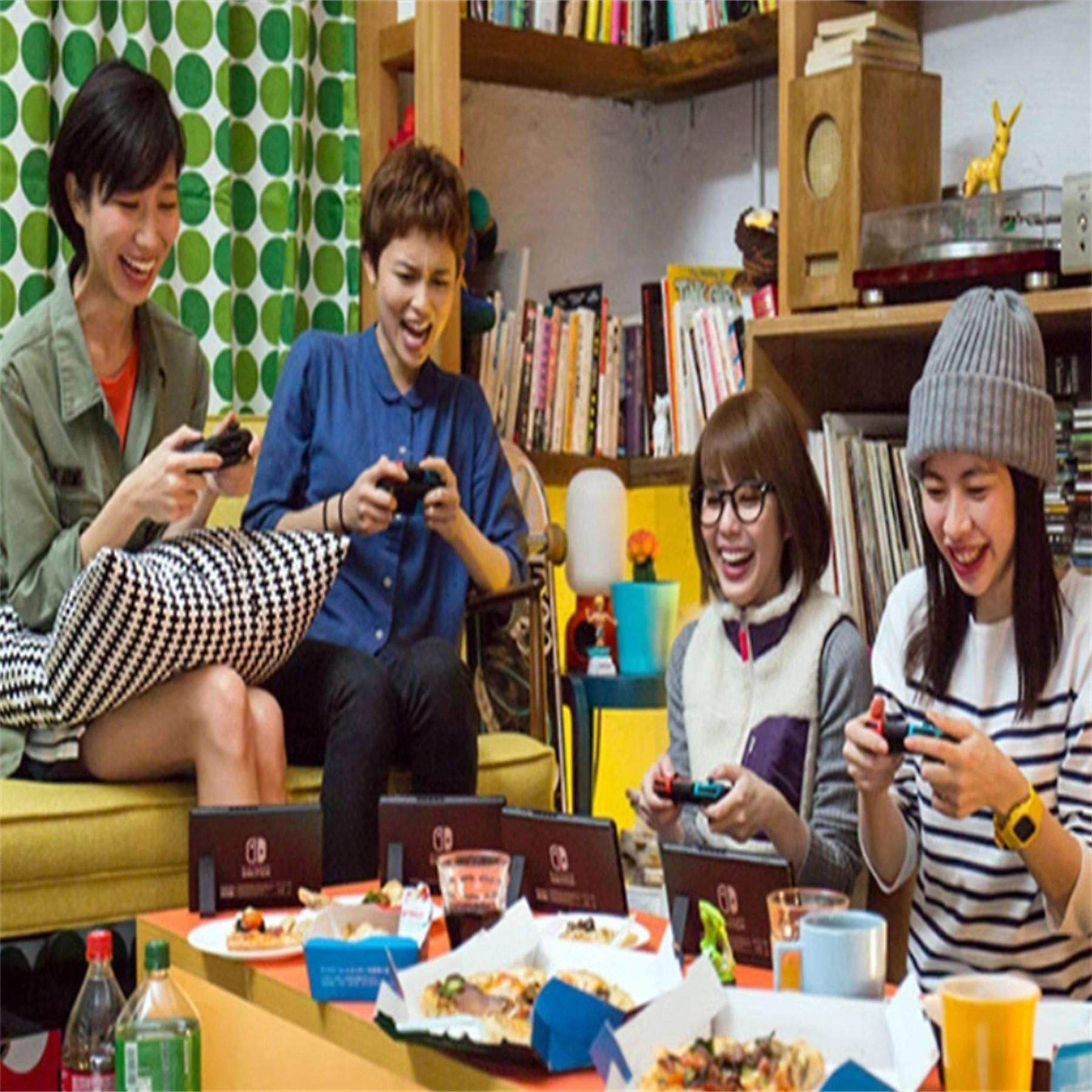 Vier Frauen lachen, während sie mit Controllern Videospiele spielen. Pizza und Getränke stehen auf dem Tisch vor ihnen.