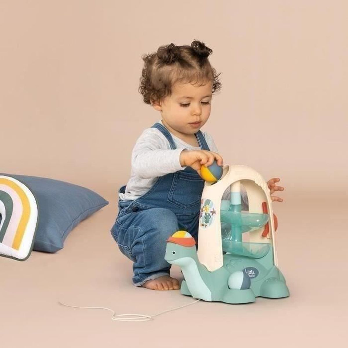 Un bambino piccolo con tuta di jeans gioca con un giocattolo a forma di tartaruga, facendovi cadere una palla. Un cuscino e una decorazione arcobaleno sullo sfondo.