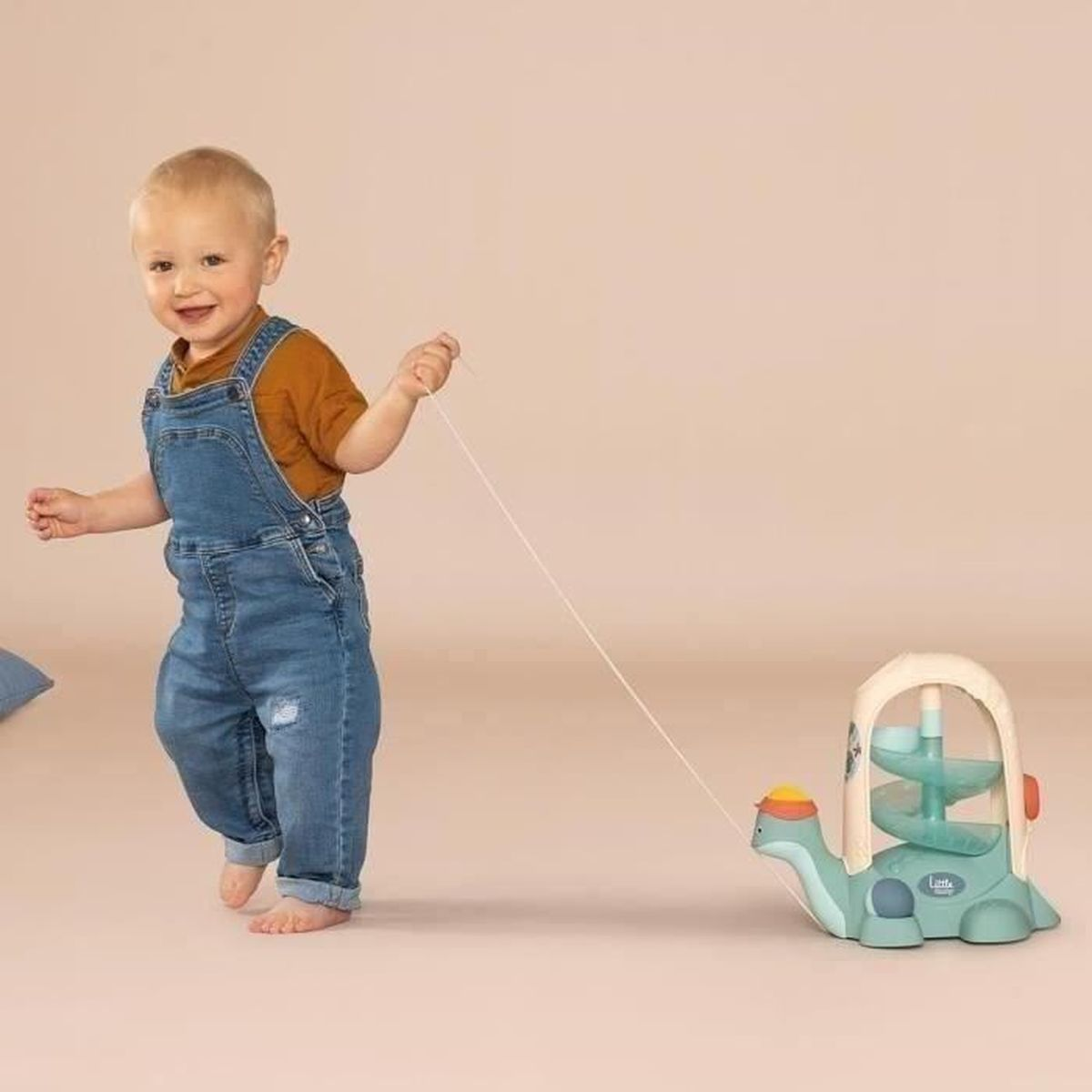 Un bambino in tuta di jeans tira un giocattolo a forma di tartaruga con una corda. Il giocattolo è verde acqua e bianco. Il bambino sorride.