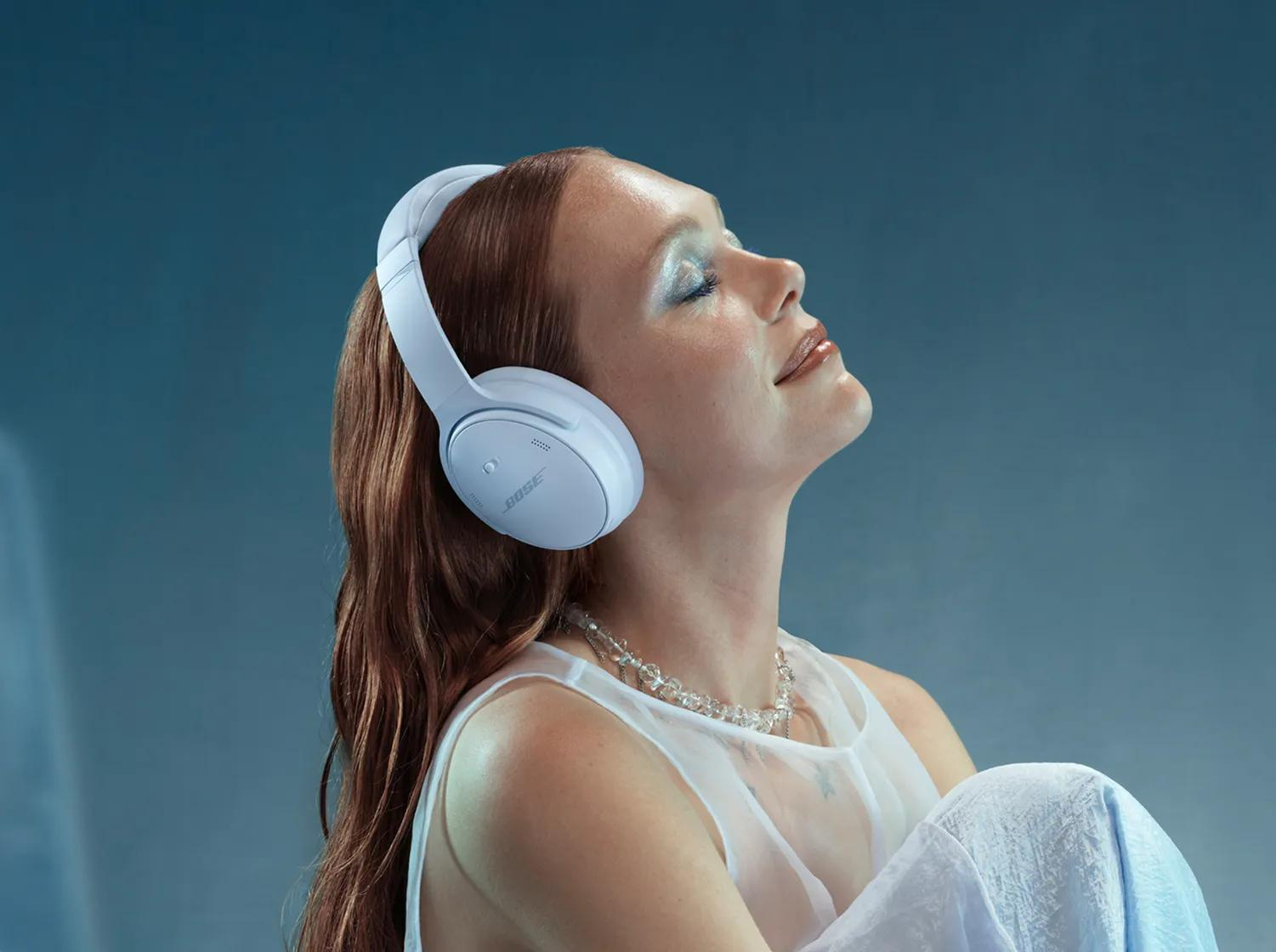 Una donna con i capelli rossi indossa cuffie bianche, una collana e un top bianco, che guarda in alto. Sfondo blu.