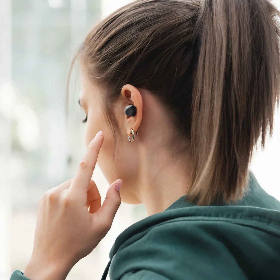Donna con un auricolare nero nell'orecchio, che lo indica. Indossa una felpa con cappuccio verde e ha una coda di cavallo. È visibile un orecchino a cerchio dorato.