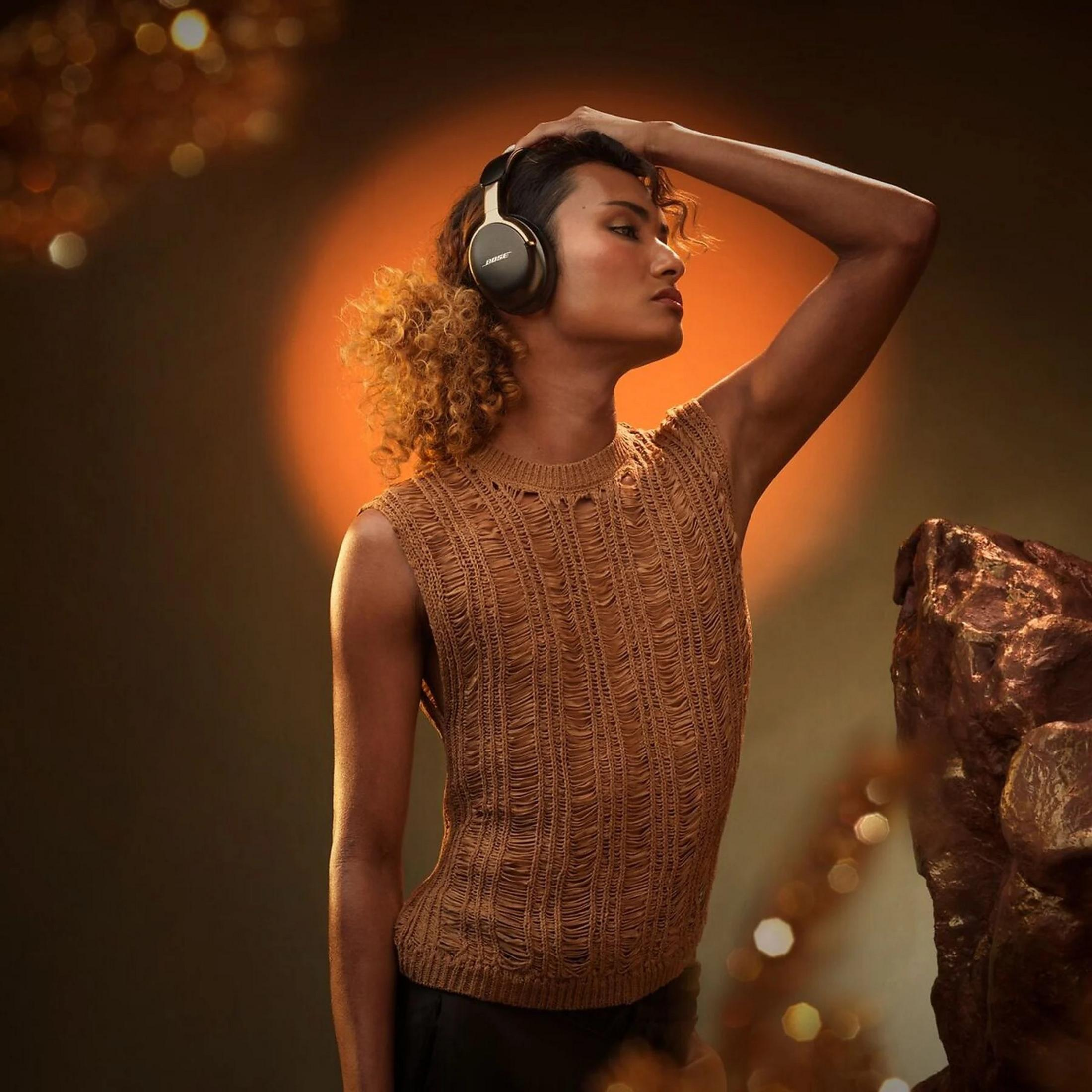 Una persona con auriculares y una camiseta marrón, con la mano en la cabeza, sobre un fondo naranja. Son visibles los auriculares Bose.