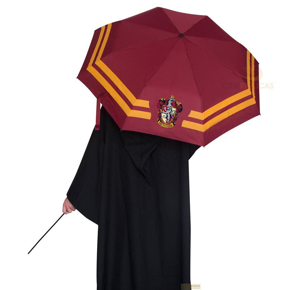 Una persona con una túnica negra sostiene una varita y un paraguas granate abierto con rayas doradas y un escudo de Gryffindor.