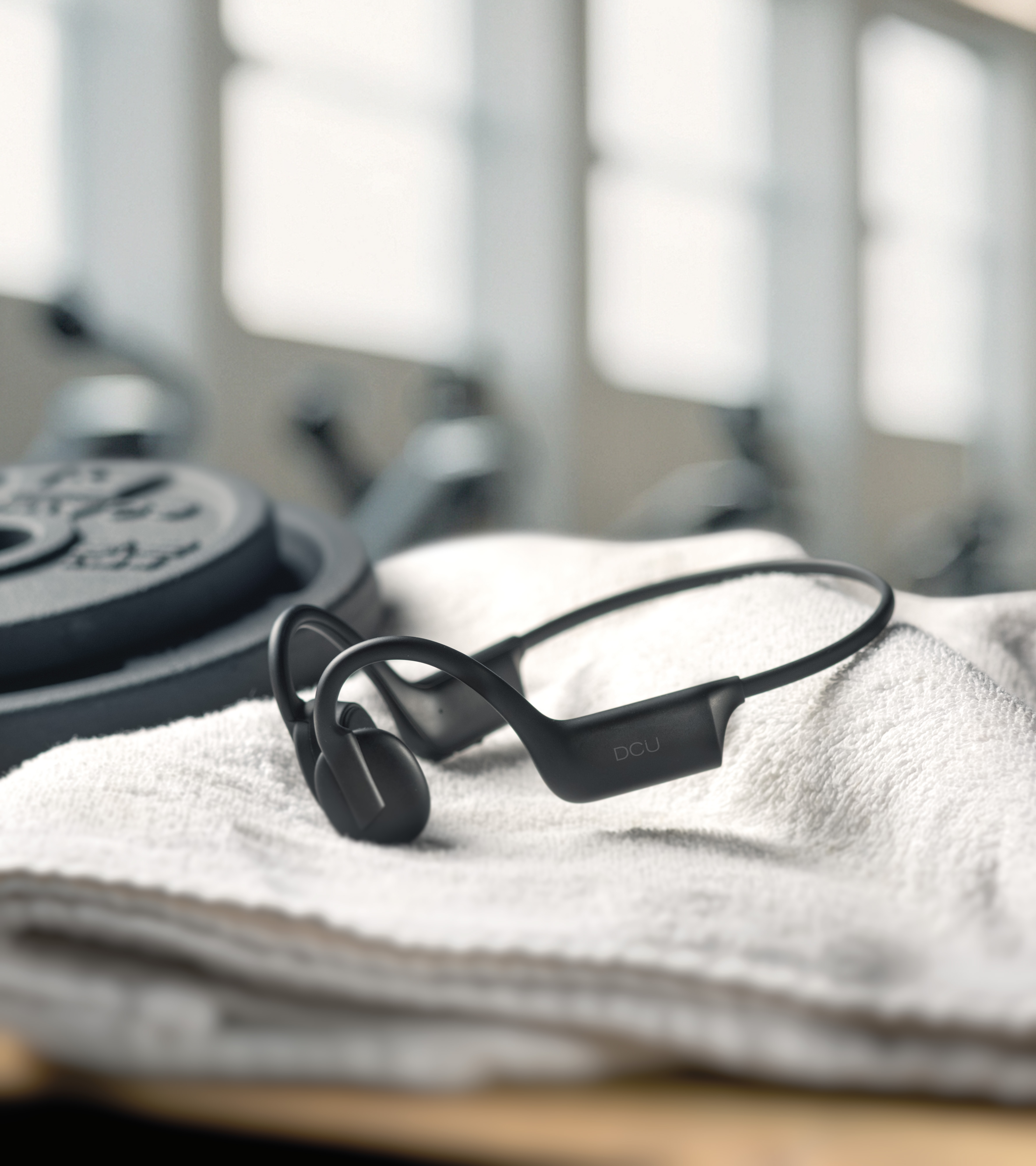 Auriculares deportivos negros sobre una toalla blanca. Discos de pesas y fondo de gimnasio borroso.