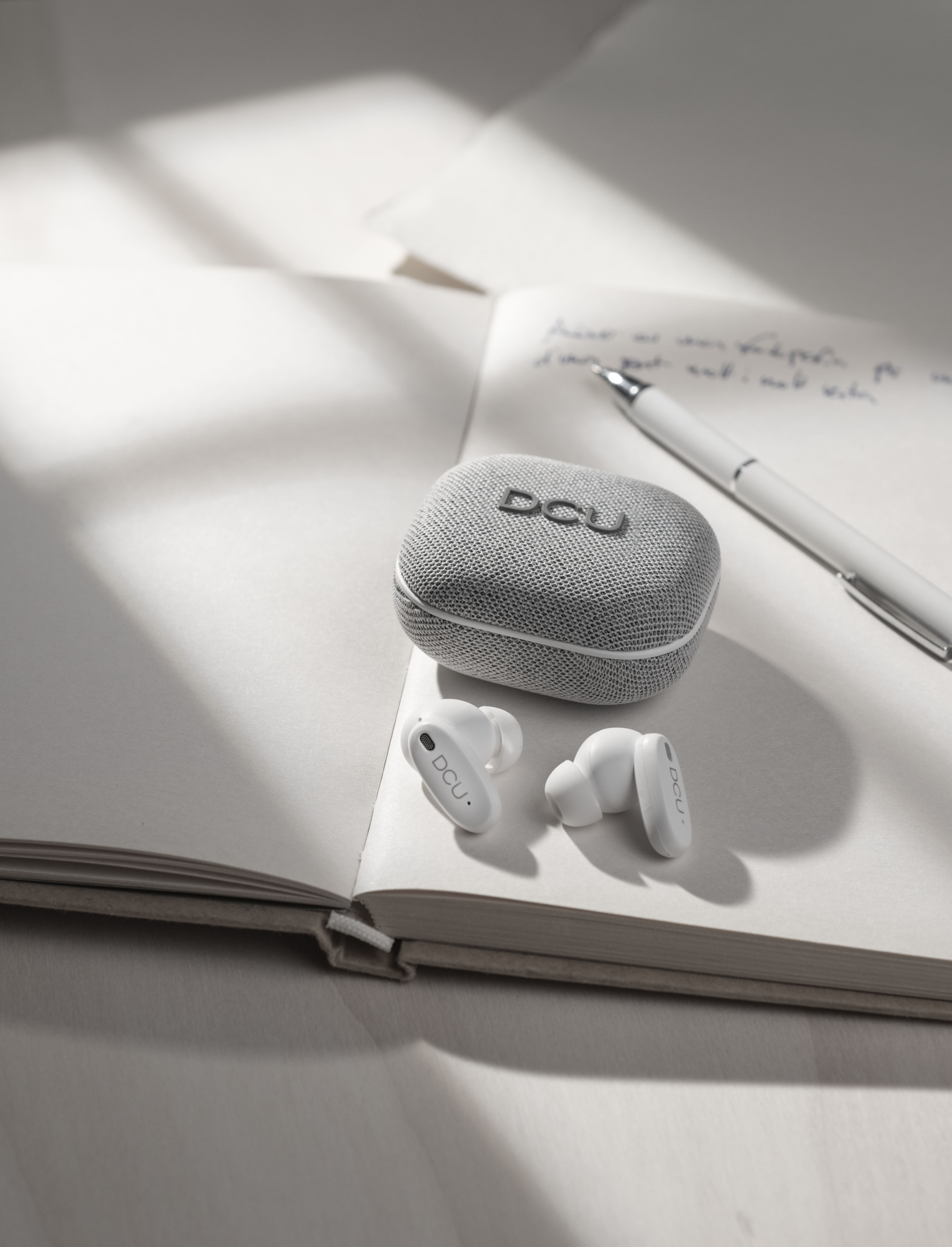 Weiße Ohrhörer, Etui und Stift auf einem offenen Notizbuch. Schatten spielen auf der Seite. Das Etui sagt DCU.