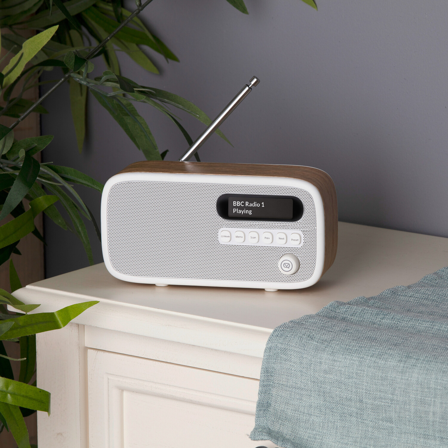 Ein Holzradio steht auf einem weißen Tisch. Ein Bildschirm zeigt 'BBC Radio 1 Playing'. Grüne Blätter links und ein blaues Tuch rechts.