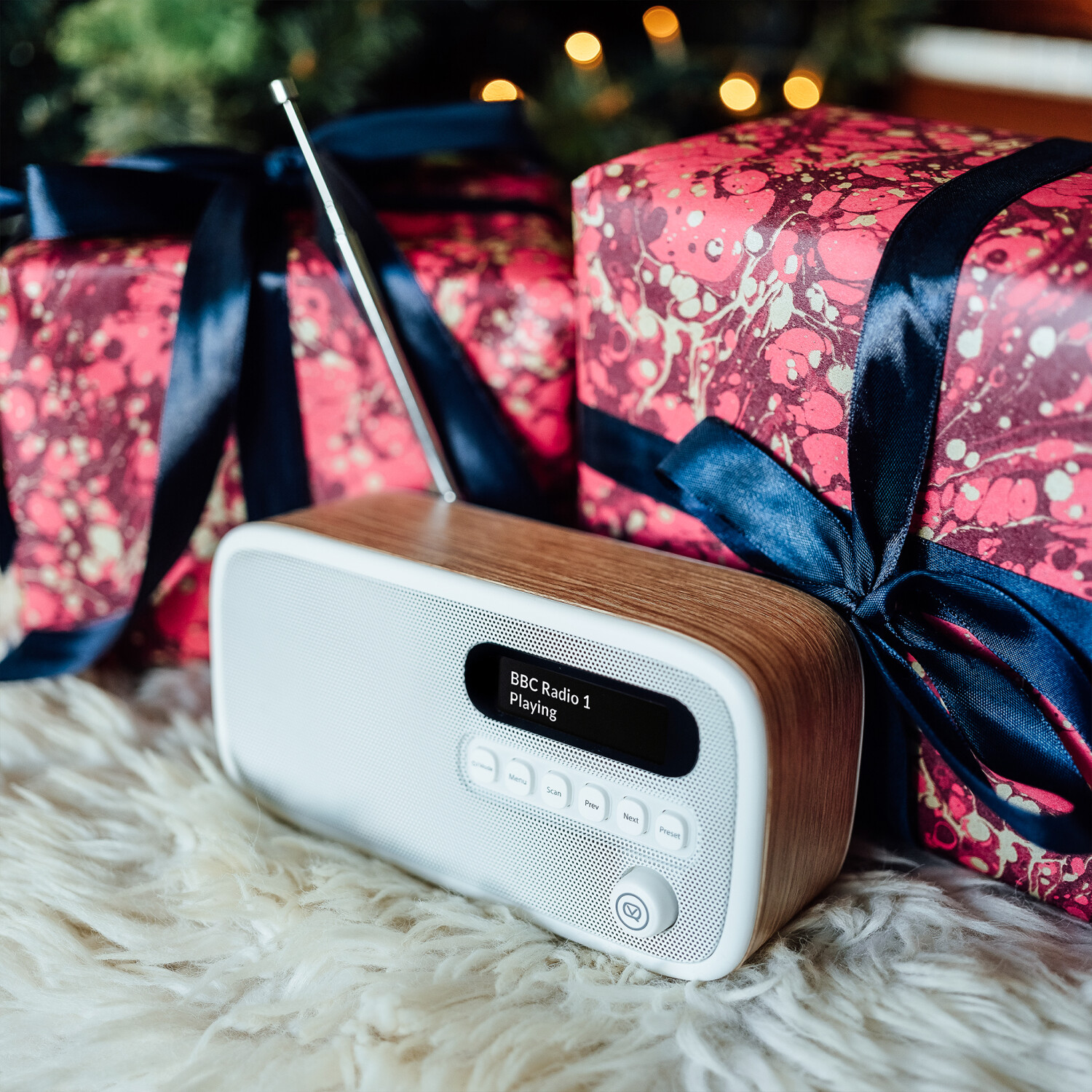 Ein weißes und holzfarbenes Radio mit der Anzeige 'BBC Radio 1 Playing' steht auf Fell. Geschenkverpackungen mit dunkelblauen Bändern im Hintergrund.