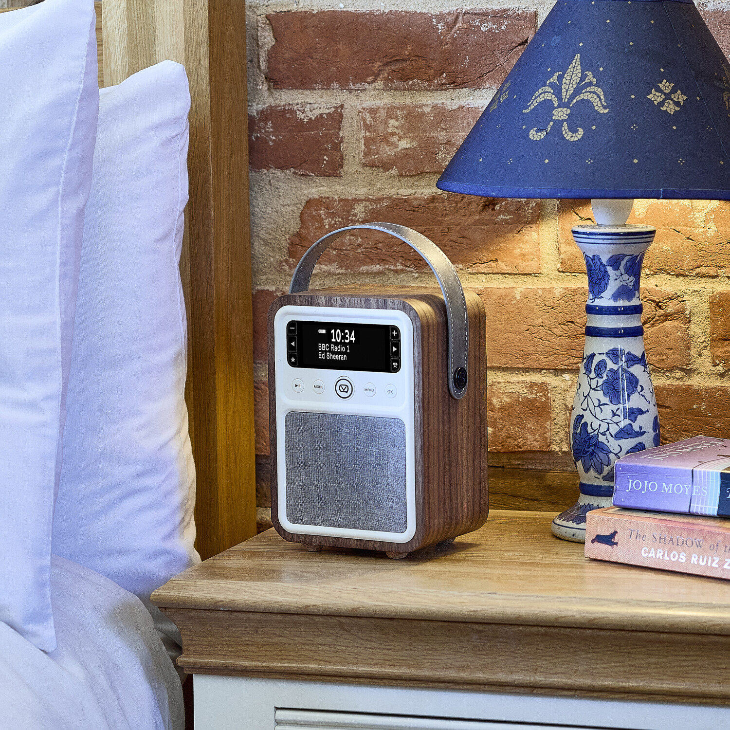 Ein Holzradio auf einem Nachttisch neben einer Lampe und Büchern. Links ein Bett. Ziegelmauer im Hintergrund.