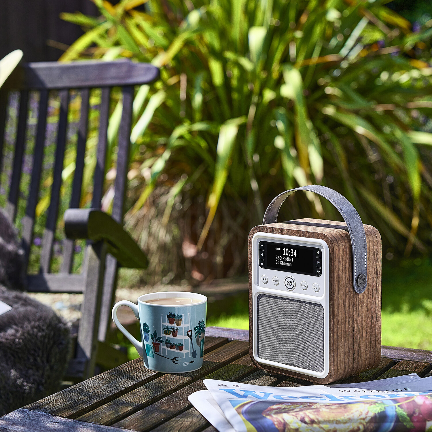 Ein Holzradio mit Griff steht auf einem Holztisch. Eine Tasse, eine Zeitung und ein Garten sind im Hintergrund. Der Bildschirm des Radios zeigt 10:34 Uhr.