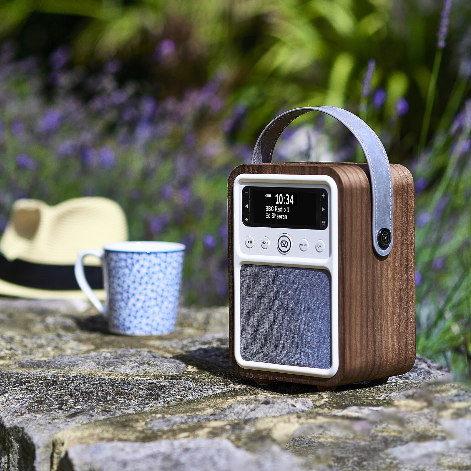 Ein Holzradio mit Griff steht auf einer Steinmauer, neben einer Tasse und einem Hut, mit verschwommenem Gartenhintergrund.