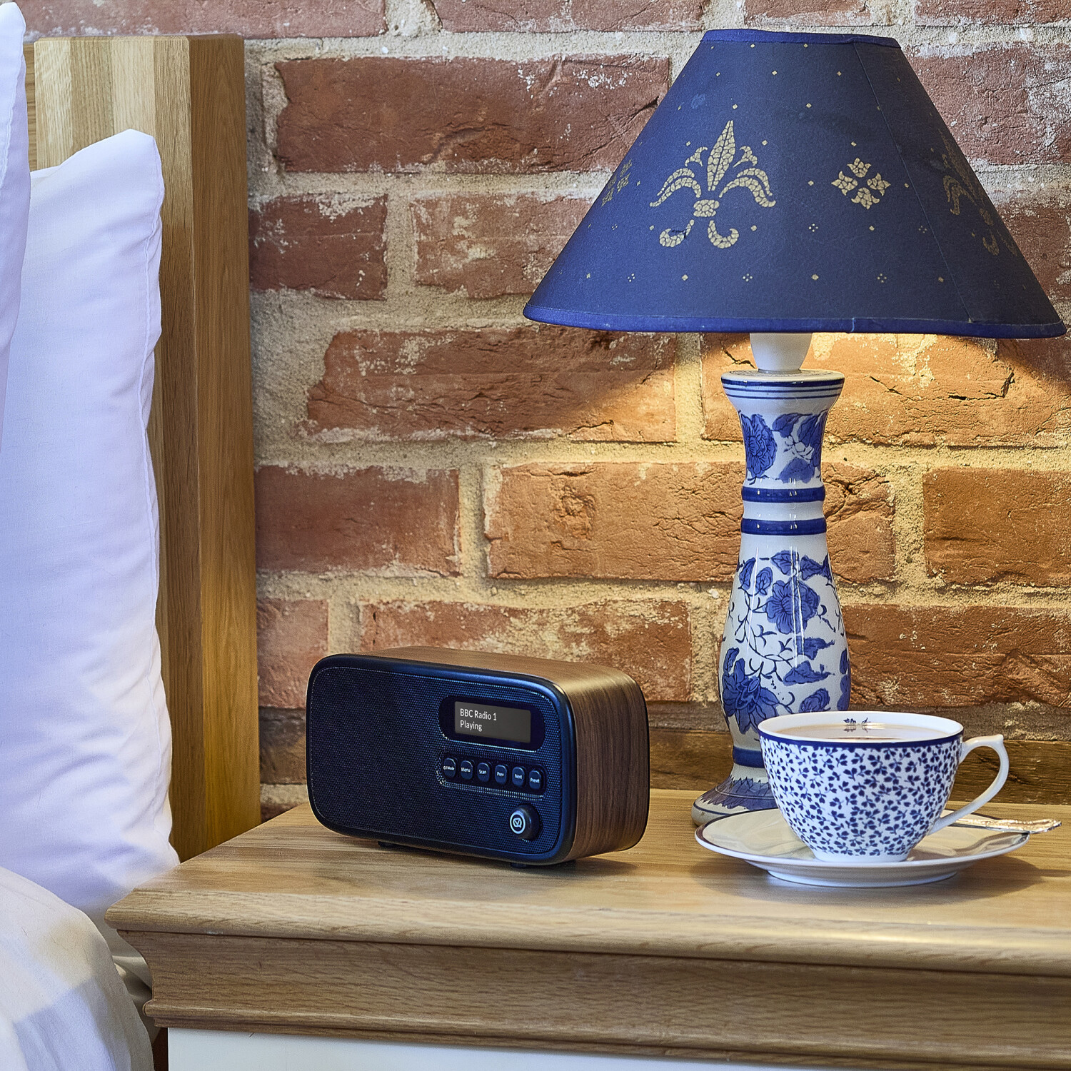 Ein Nachttisch mit Radio, Lampe und Teetasse. Ein weißes Kissen und ein Holzbettrahmen befinden sich links. Eine Backsteinmauer dient als Hintergrund.