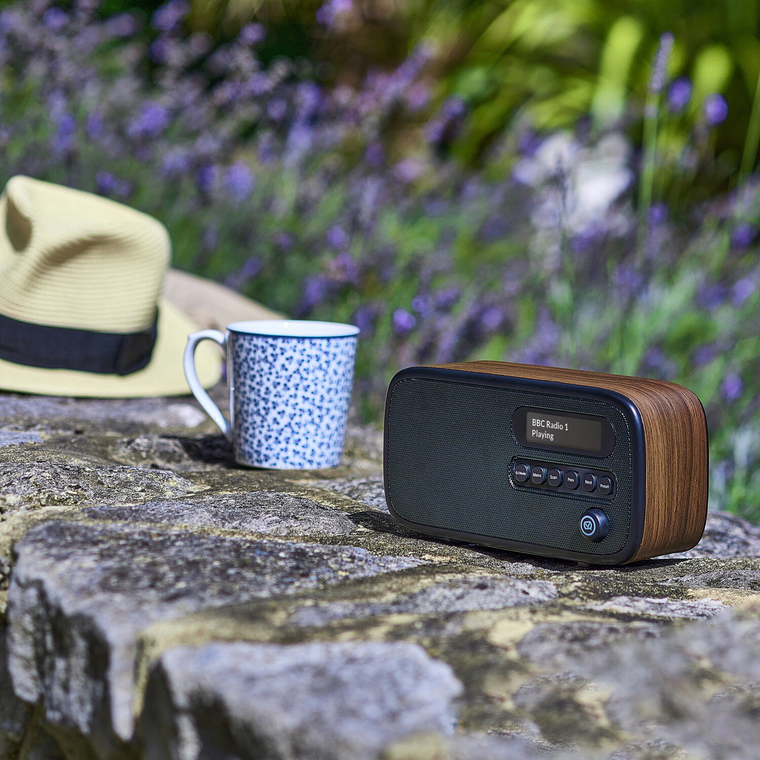 Ein Holzradio mit BBC Radio 1 steht auf einer Steinmauer, neben einer Tasse und einem Strohhut, mit lila Blumen im Hintergrund.