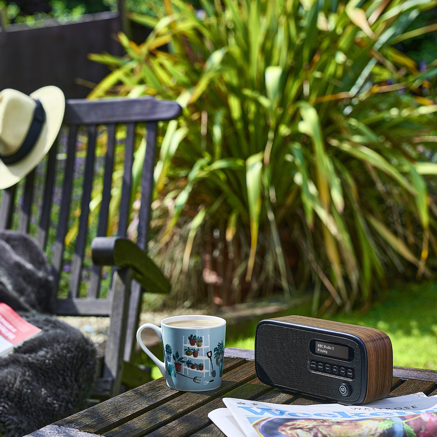 Ein Holzradio steht auf einem Tisch mit einer Kaffeetasse und einer Zeitung. Ein Hut auf einem Stuhl im Hintergrund, eine Gartenszene.