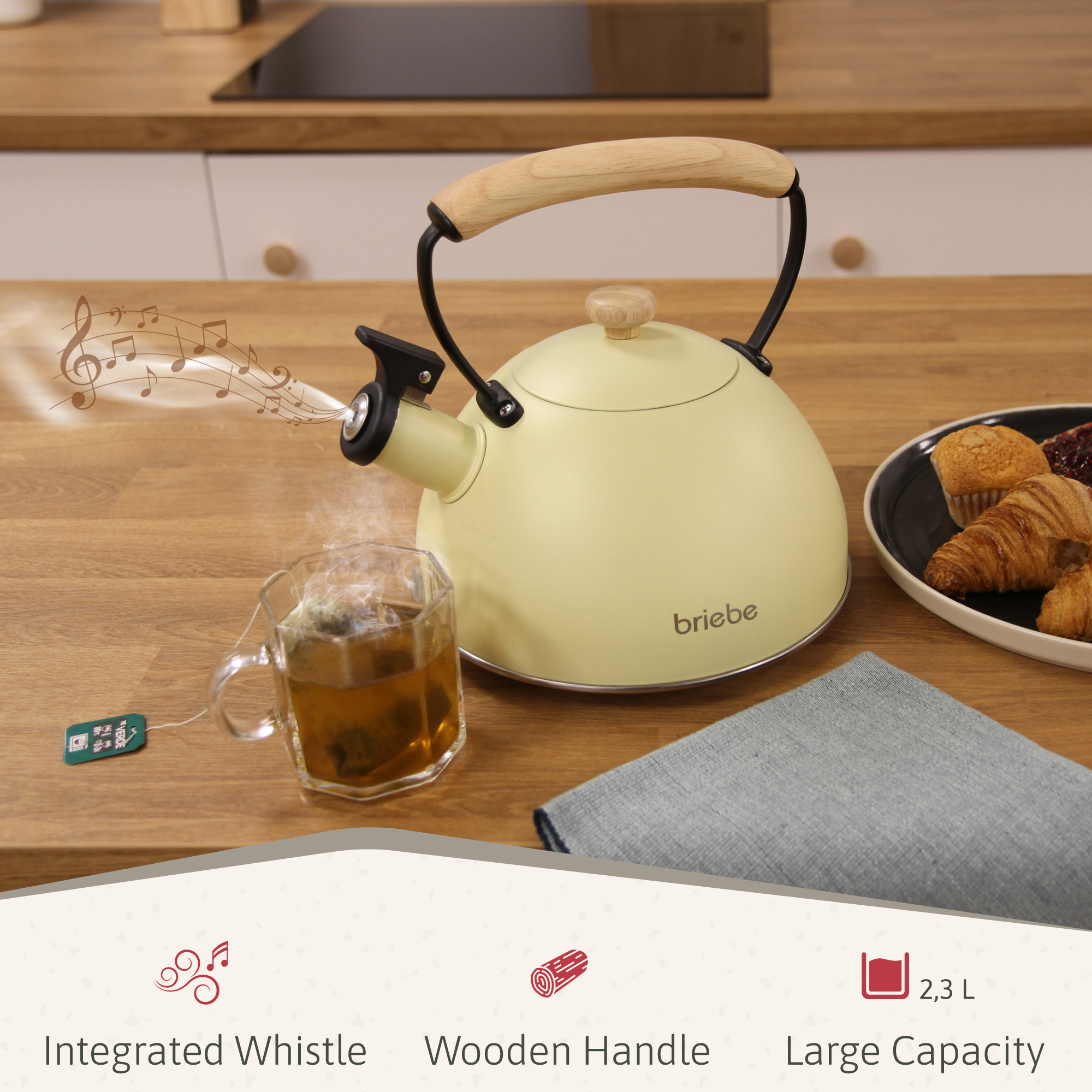 Eine cremefarbene Teekanne mit Holzgriff pfeift. Eine Tasse Tee und Gebäck stehen auf einem Holztisch.