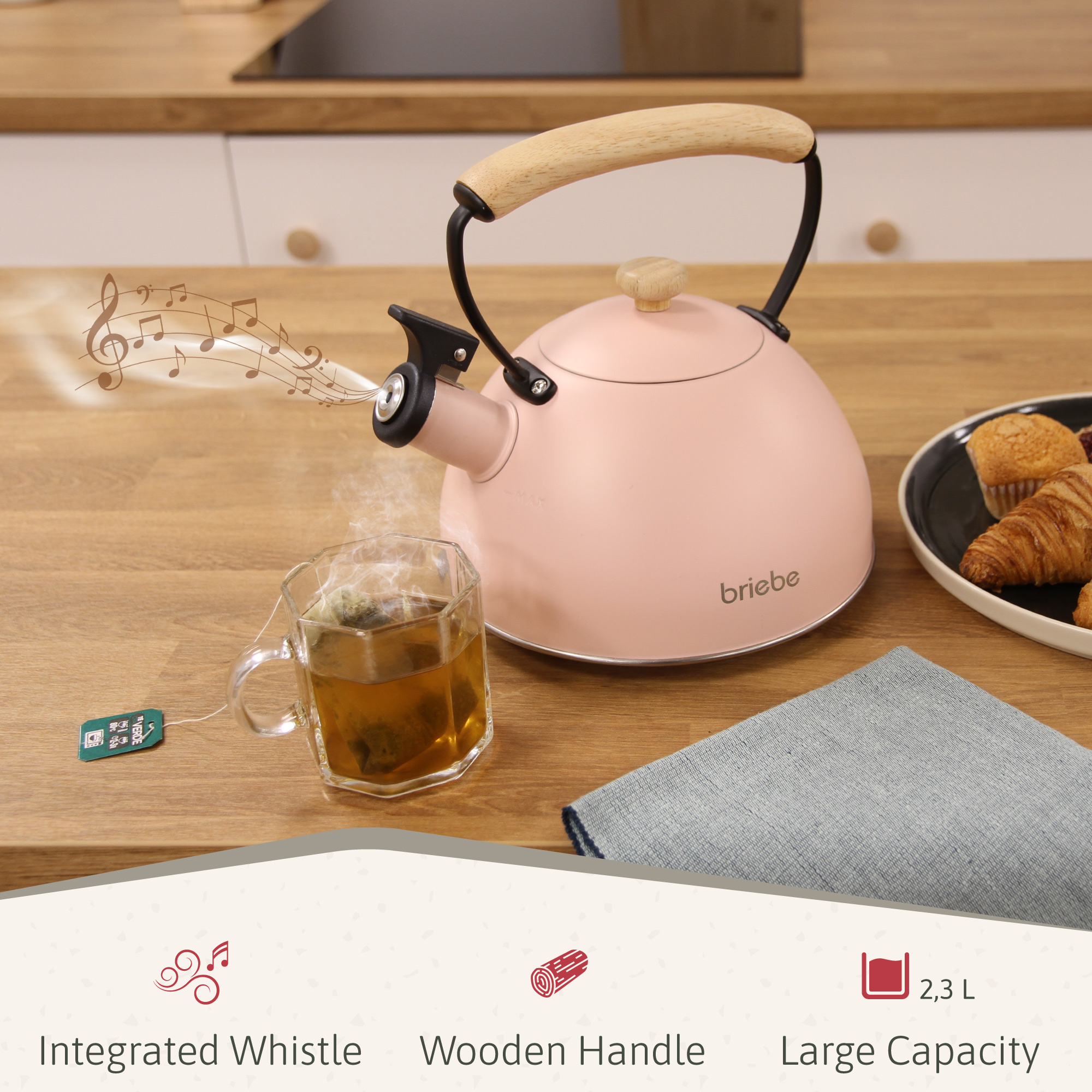 Tetera rosa con asa de madera sobre mesa de madera. Una taza de té humea junto a ella, con notas musicales. Magdalenas y servilleta cerca.
