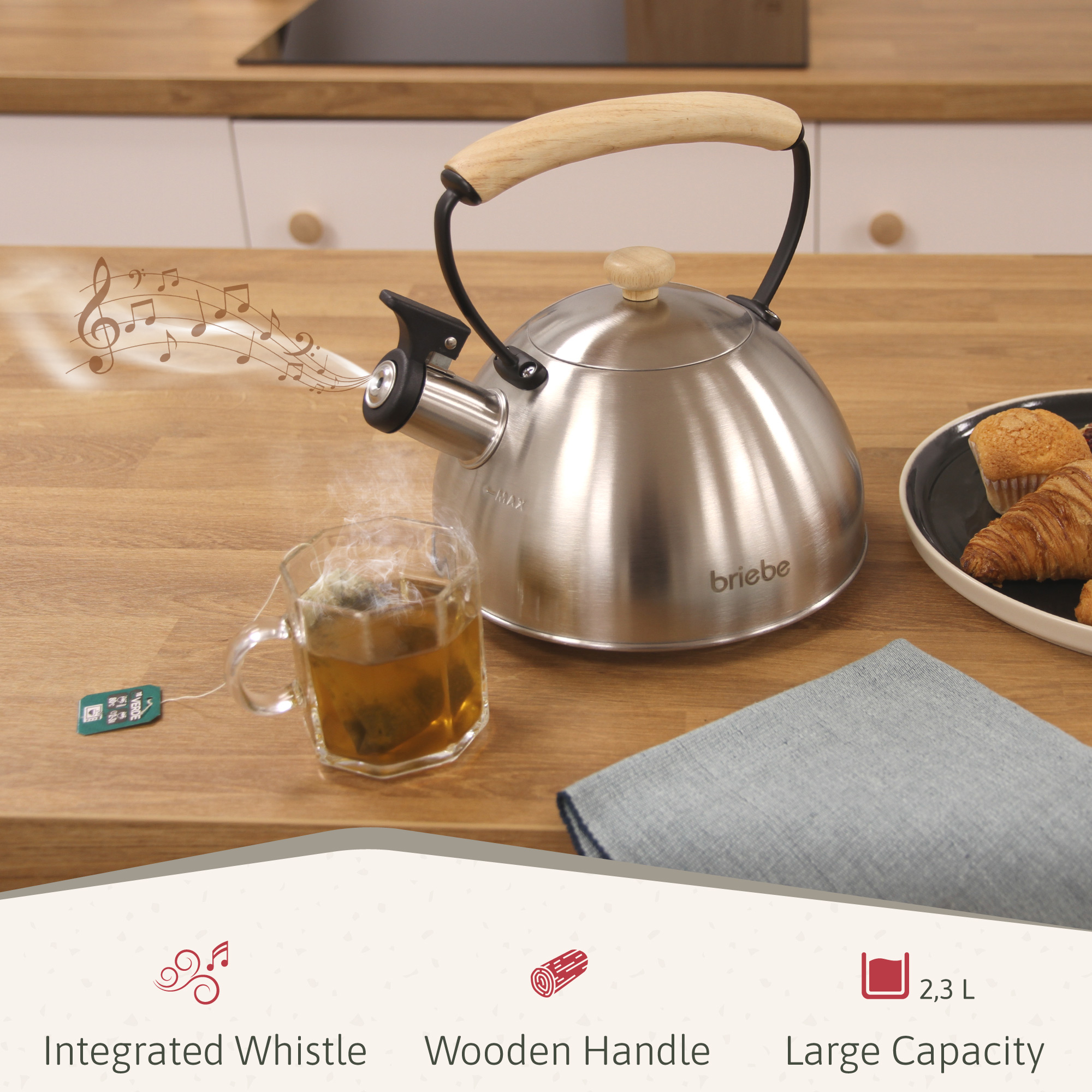 Ein Edelstahlkessel mit Holzgriff dampft, eine Teetasse mit Tee und ein Teller mit Gebäck auf einem Holztisch.