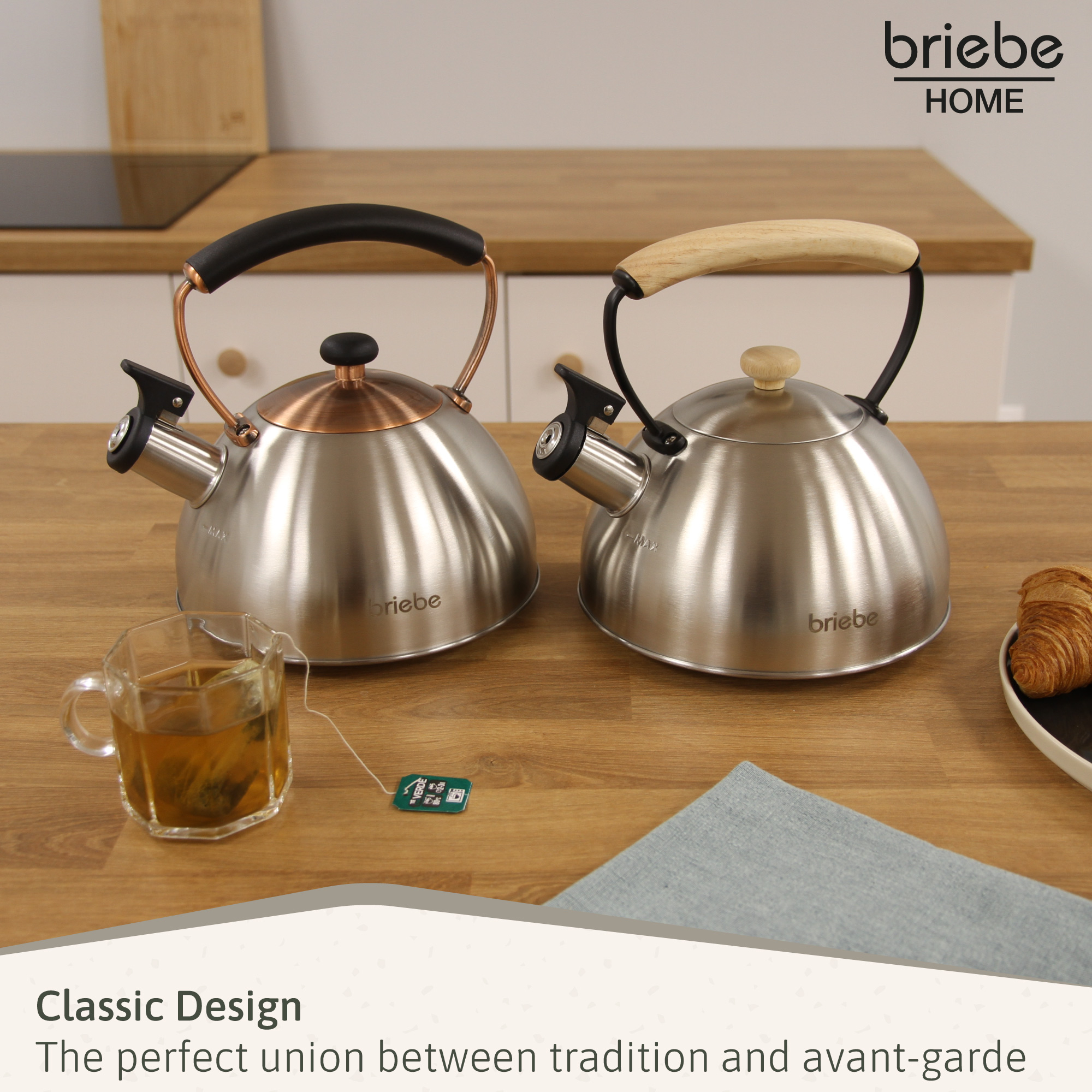 Zwei glänzende Kessel mit schwarzen und hölzernen Griffen auf einem Holztisch. Eine Teetasse und ein Croissant sind ebenfalls vorhanden.