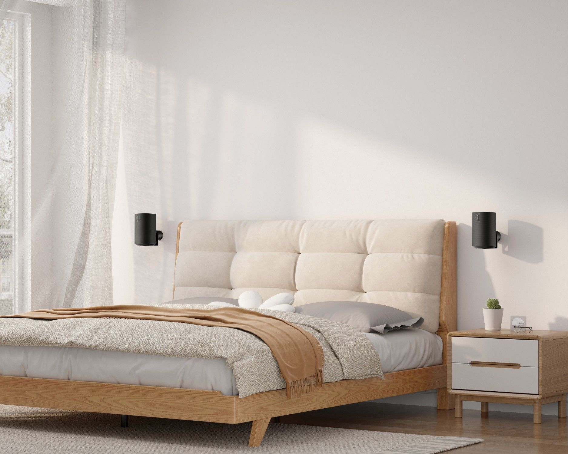 Ein Schlafzimmer mit Bett, Nachttisch und Fenster. Das Bett hat ein beiges Kopfteil und eine braune Decke. Schwarze Lautsprecher an den Wänden.