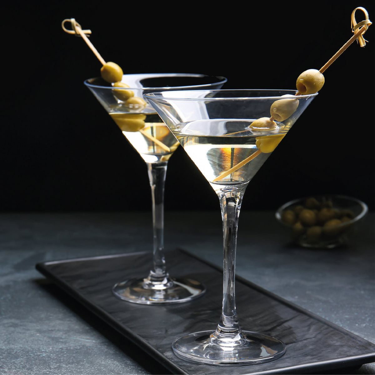 Two martini glasses with olives on a black tray. Background shows olives in a bowl and a dark surface.
