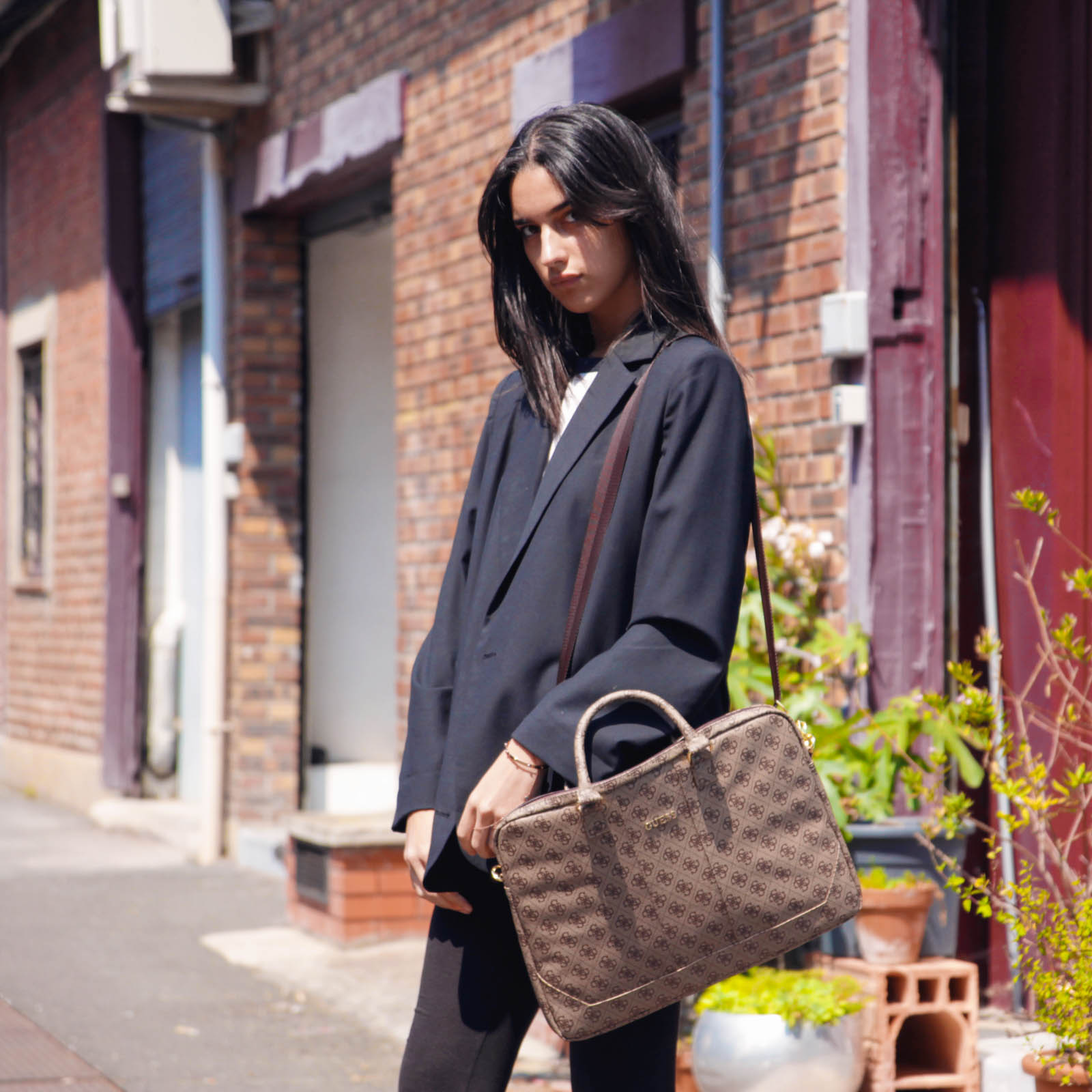Donna in blazer nero con borsa marrone in una strada con edifici in mattoni.