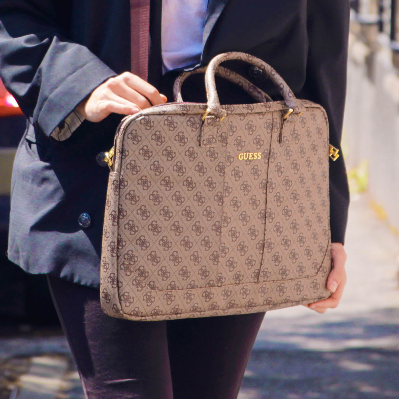 Una persona tiene in mano una borsa Guess. La borsa è color cachi con un motivo ripetuto e dettagli dorati. La persona indossa un blazer.