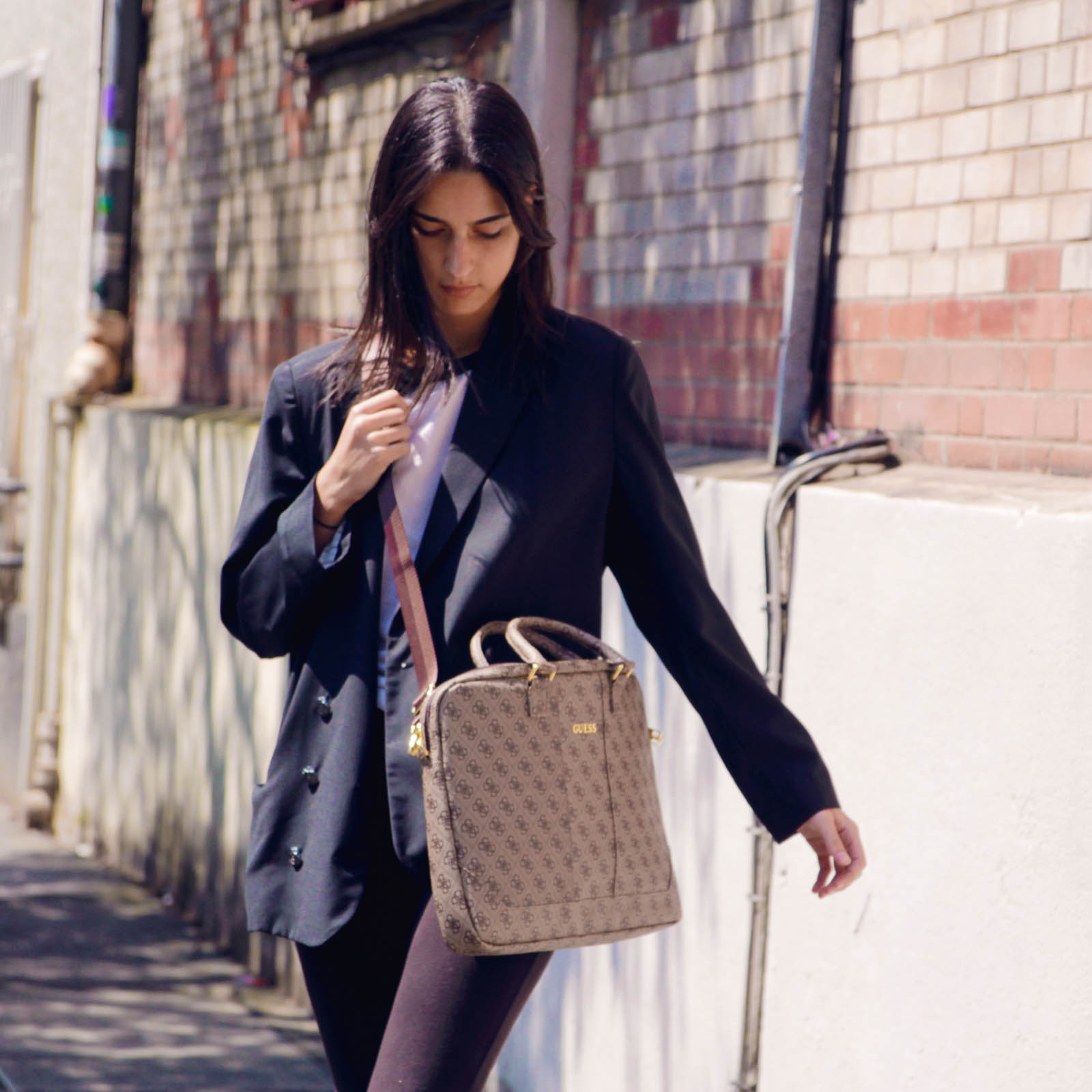 Donna in blazer nero cammina per strada, con una borsa Guess marrone. Muro di mattoni e muro bianco sullo sfondo.