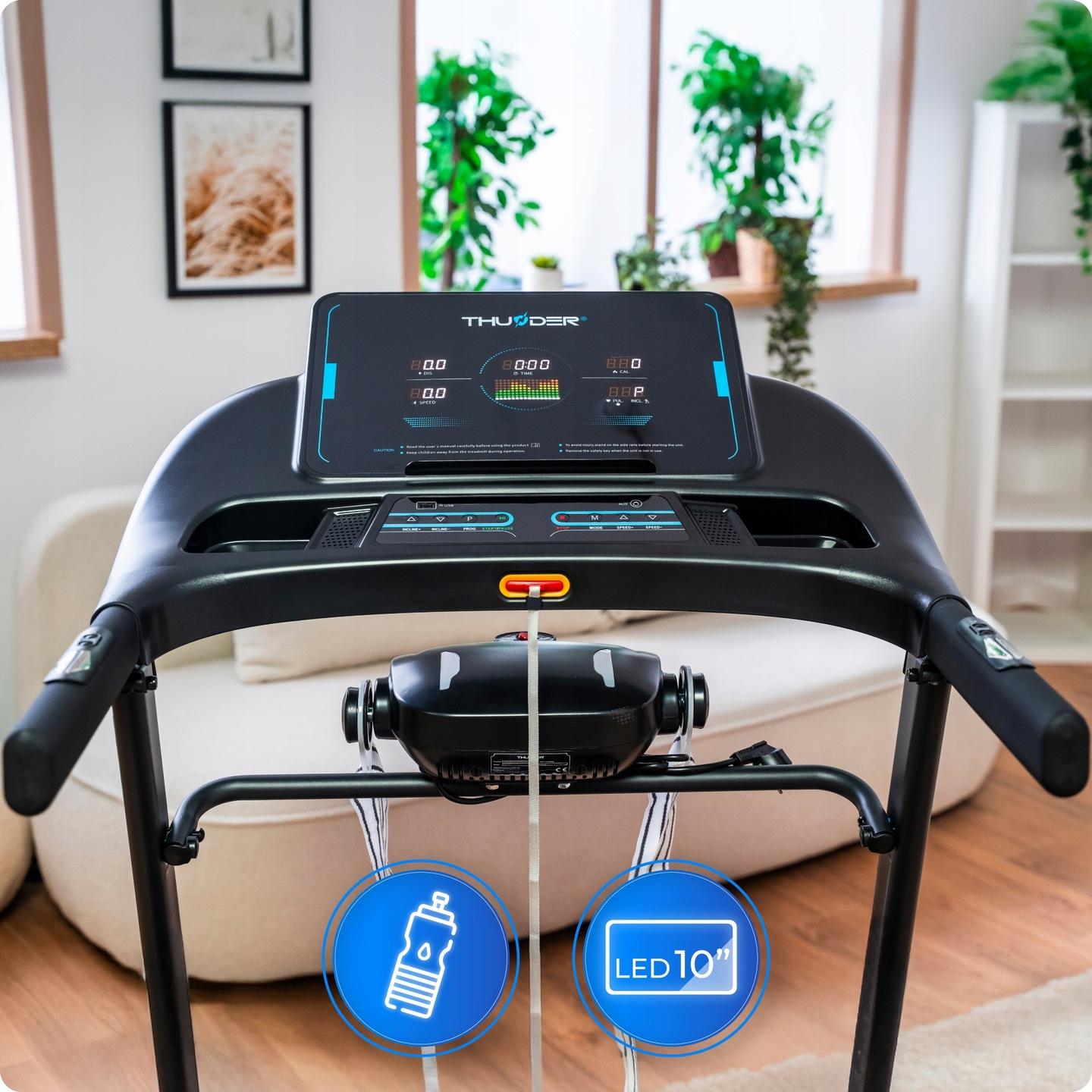 Bieżnia stoi w pokoju. Wyświetlacz pokazuje liczby. Na maszynie znajduje się butelka wody i znak LED 10".