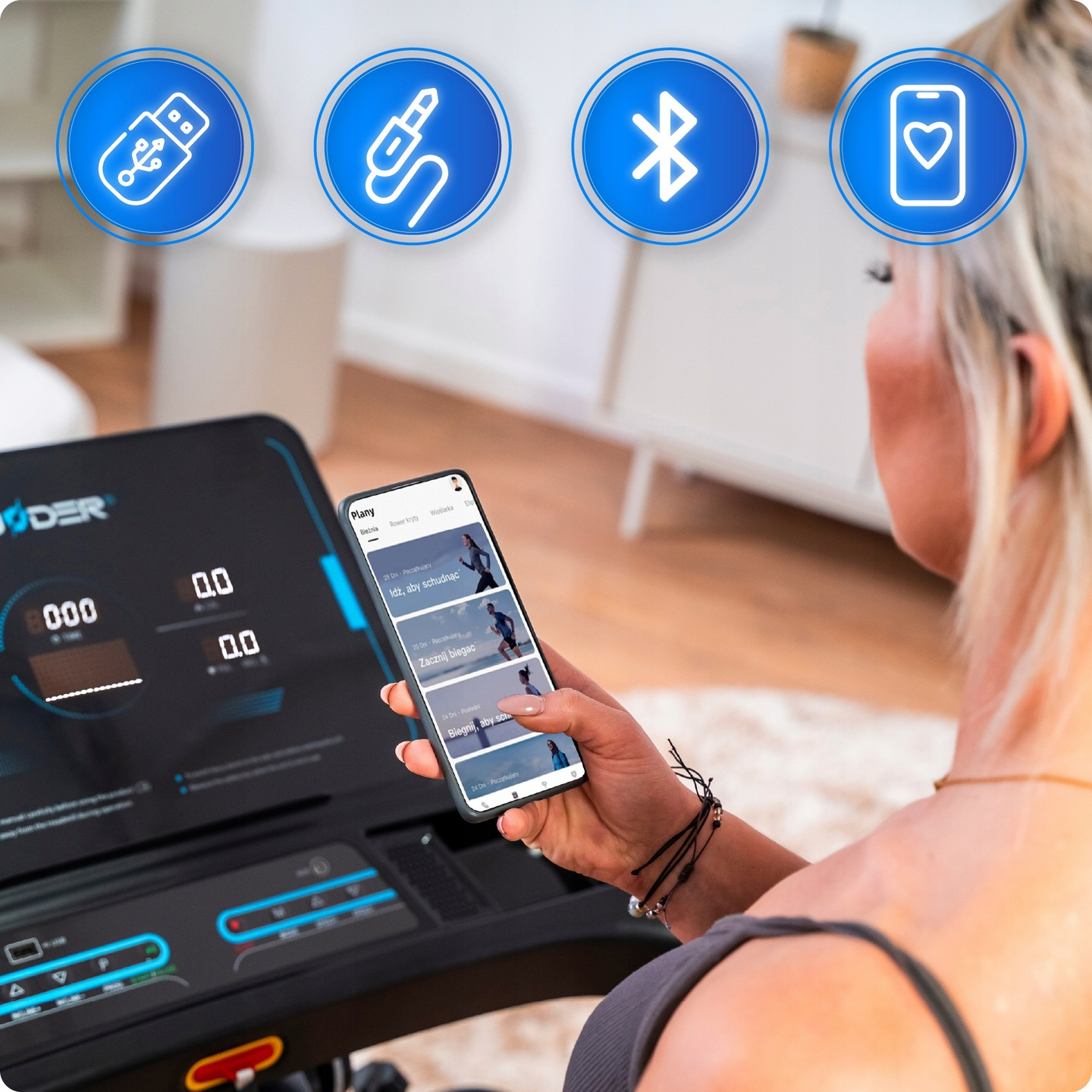 Kobieta używa smartfona z planami treningowymi obok bieżni. Widoczne ikony USB, gniazda audio, Bluetooth i serca.