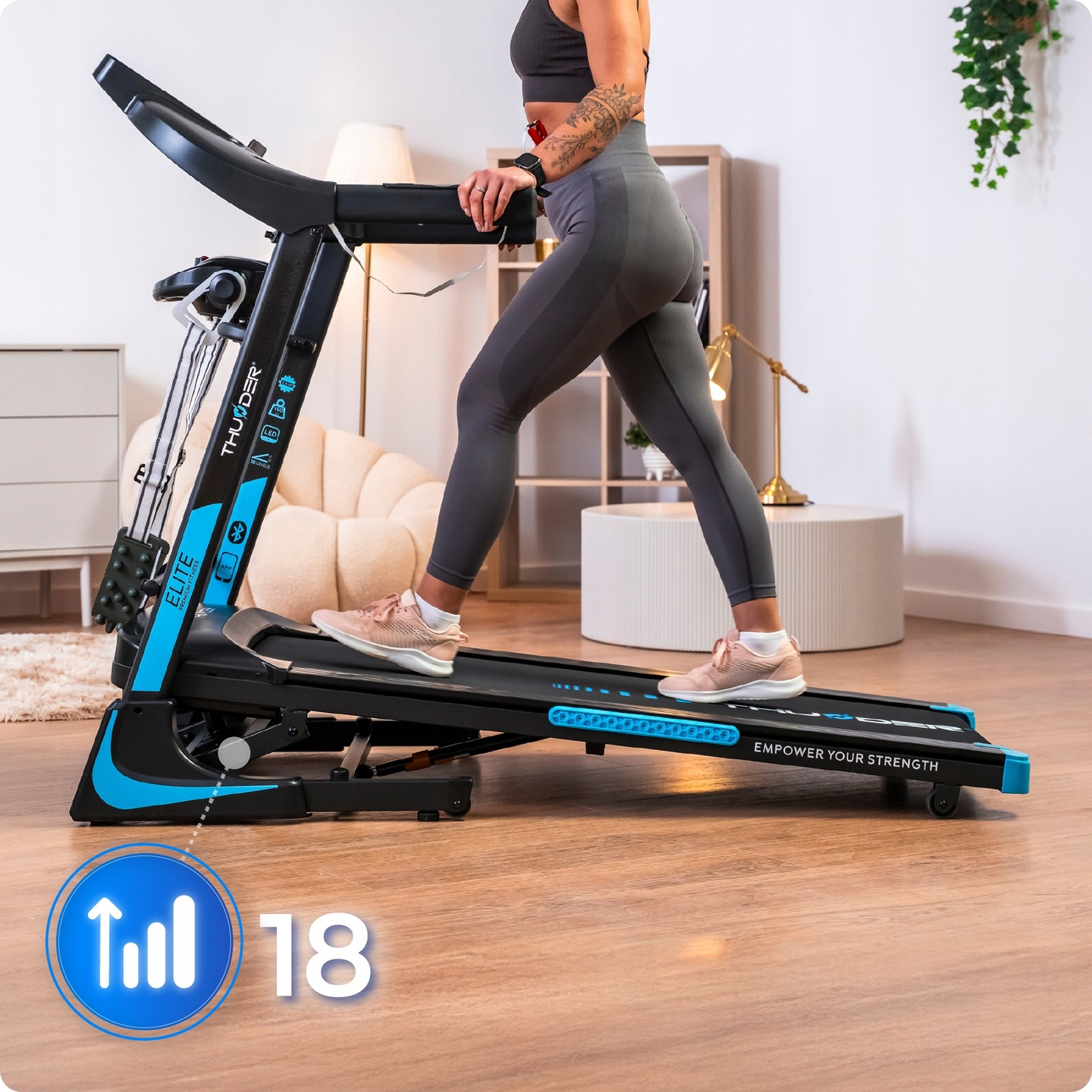 Kobieta ćwiczy na bieżni. Maszyna jest czarna i niebieska. Tło przedstawia białą ścianę i meble.