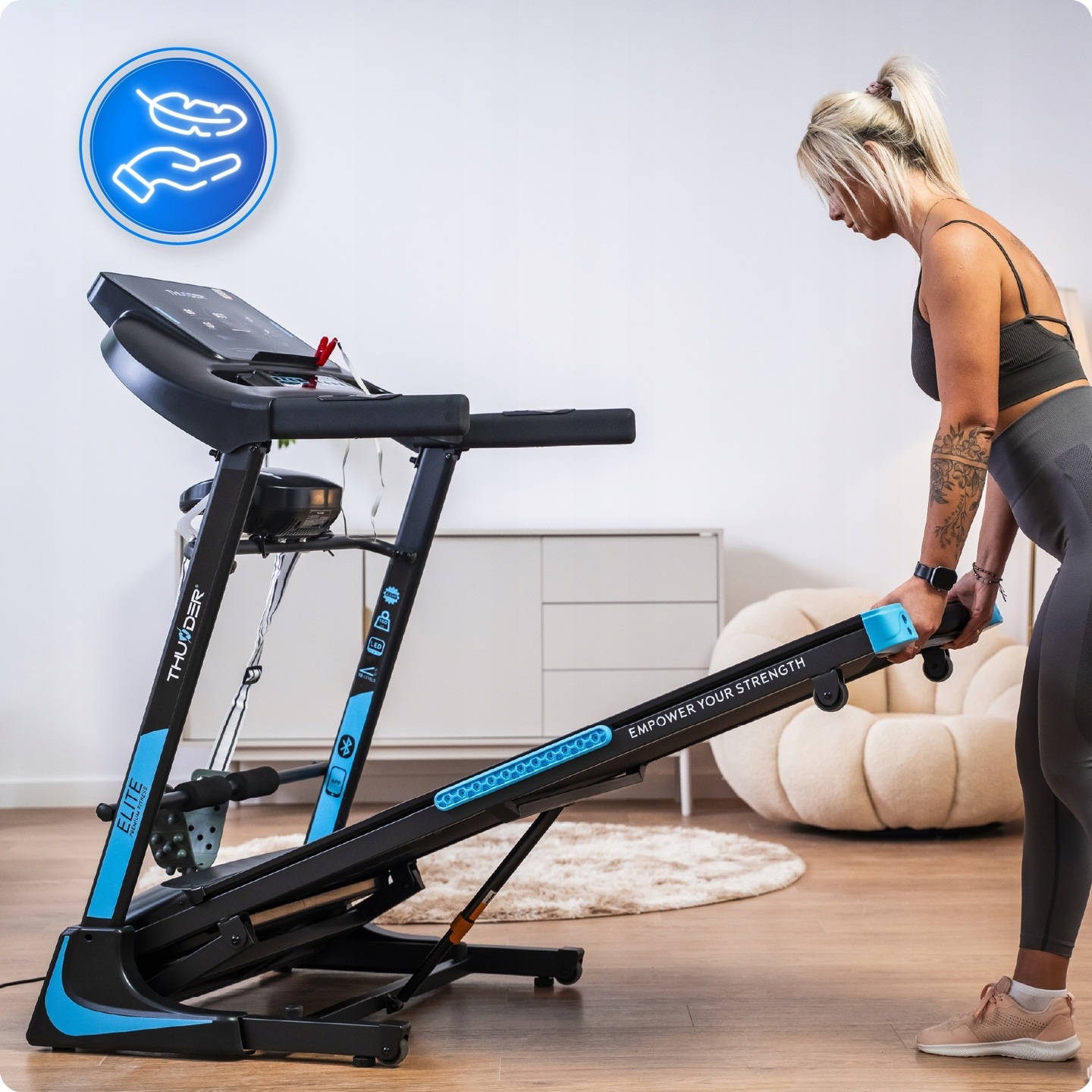 Kobieta reguluje bieżnię w pokoju. Maszyna jest czarno-niebieska. Za nią znajduje się miękki fotel i biała szafka.