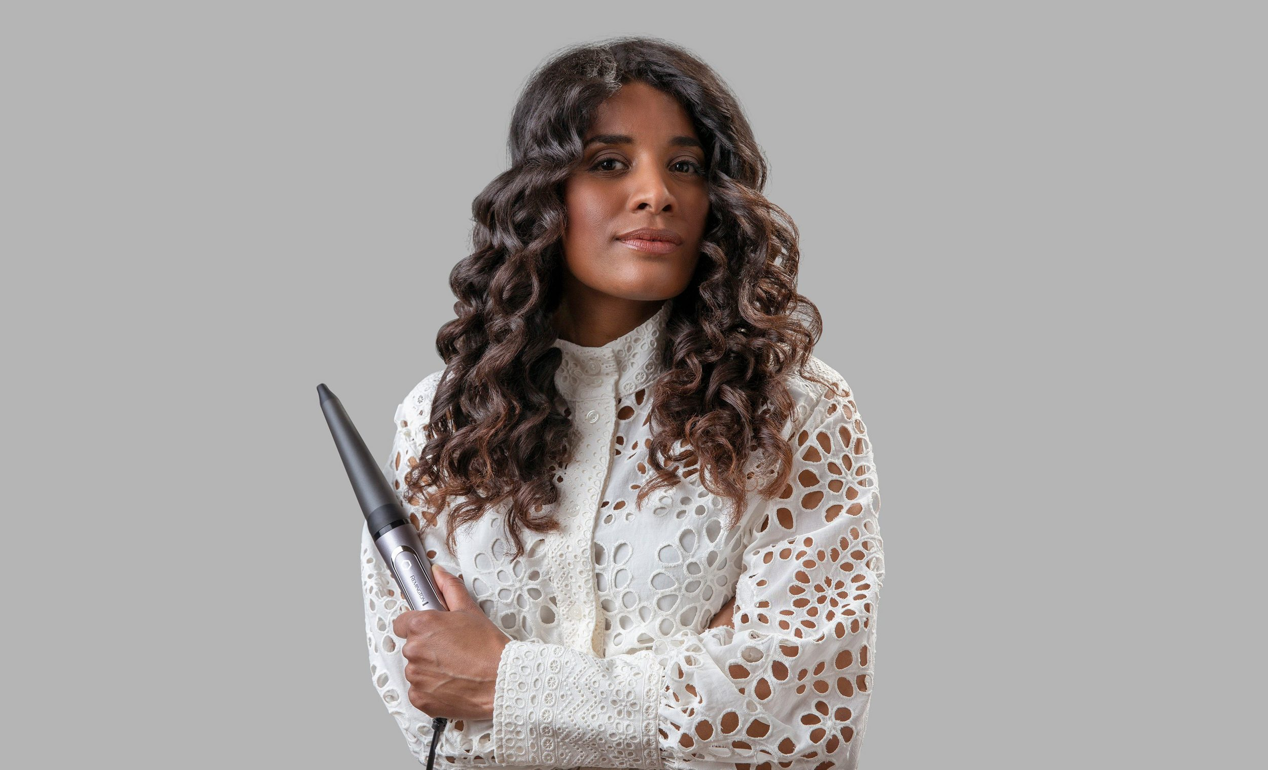 Kobieta z kręconymi włosami trzyma lokówkę. Ma na sobie białą bluzkę z wycinanymi wzorami. Szare tło.