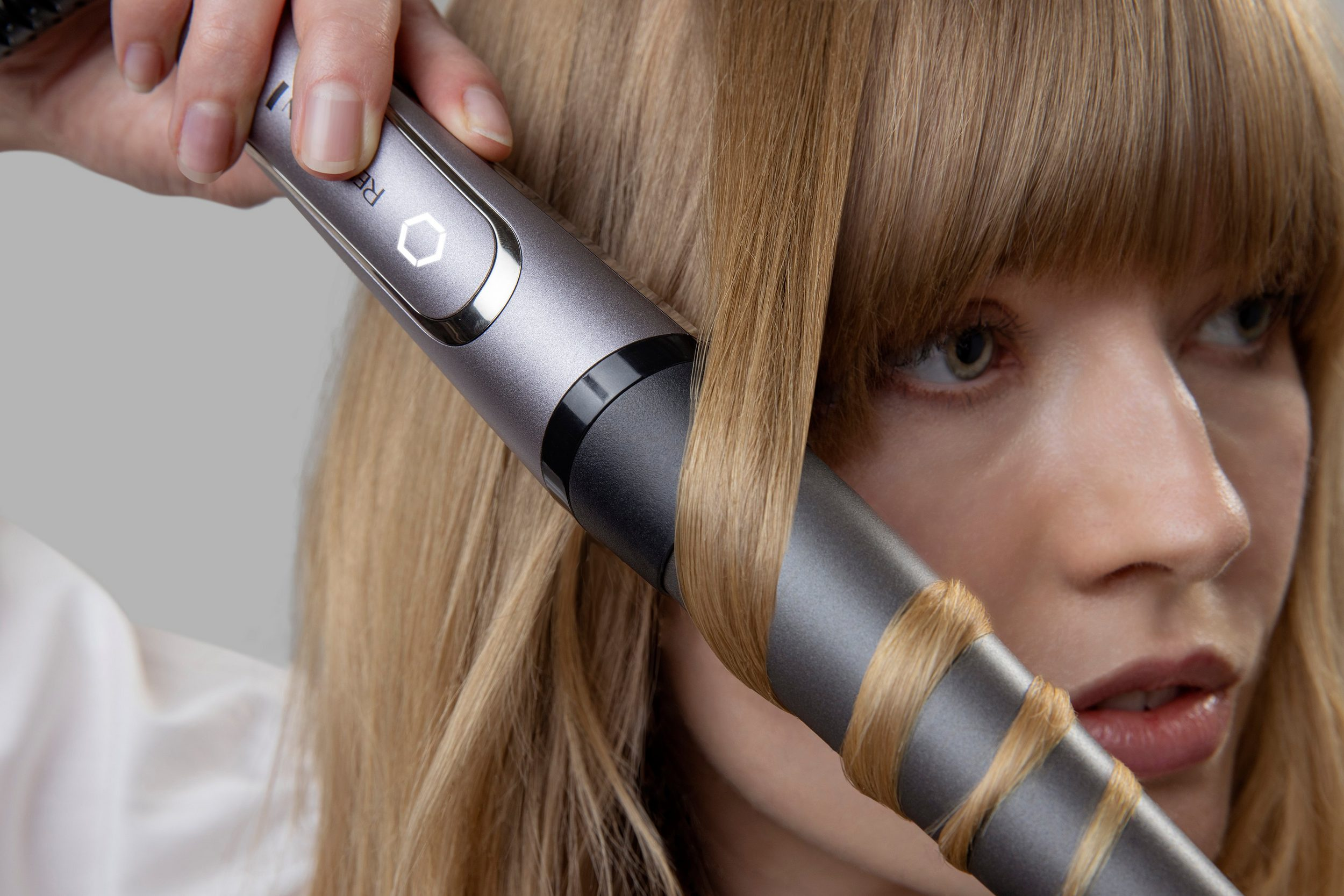 Kobieta używa lokówki na swoich blond włosach. Lokówka jest szara. Tło jest rozmyte.