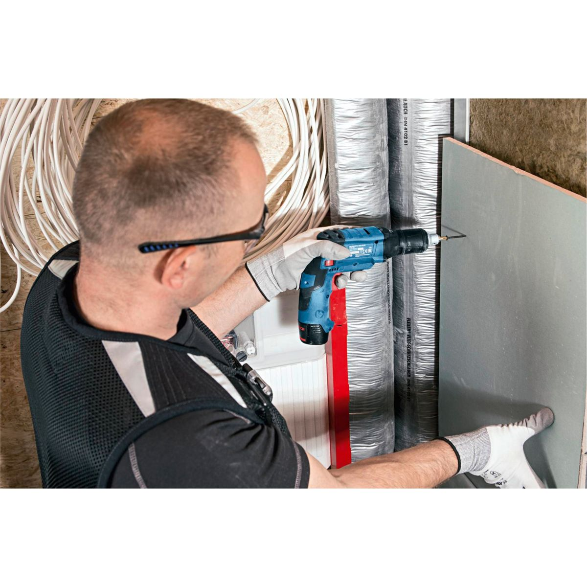 Mężczyzna wierci w ścianie niebieską wiertarką. Nosi okulary ochronne i rękawice. Przewody i izolacja w tle.