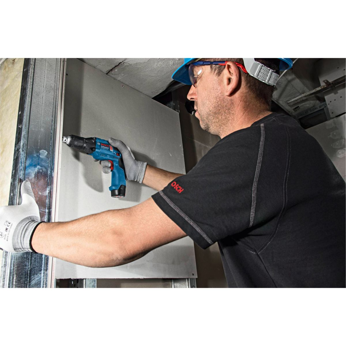 Mężczyzna w sprzęcie ochronnym używa niebieskiej wiertarki na ścianie. Ma rękawice i czarną koszulę. Scena jest w pomieszczeniu.