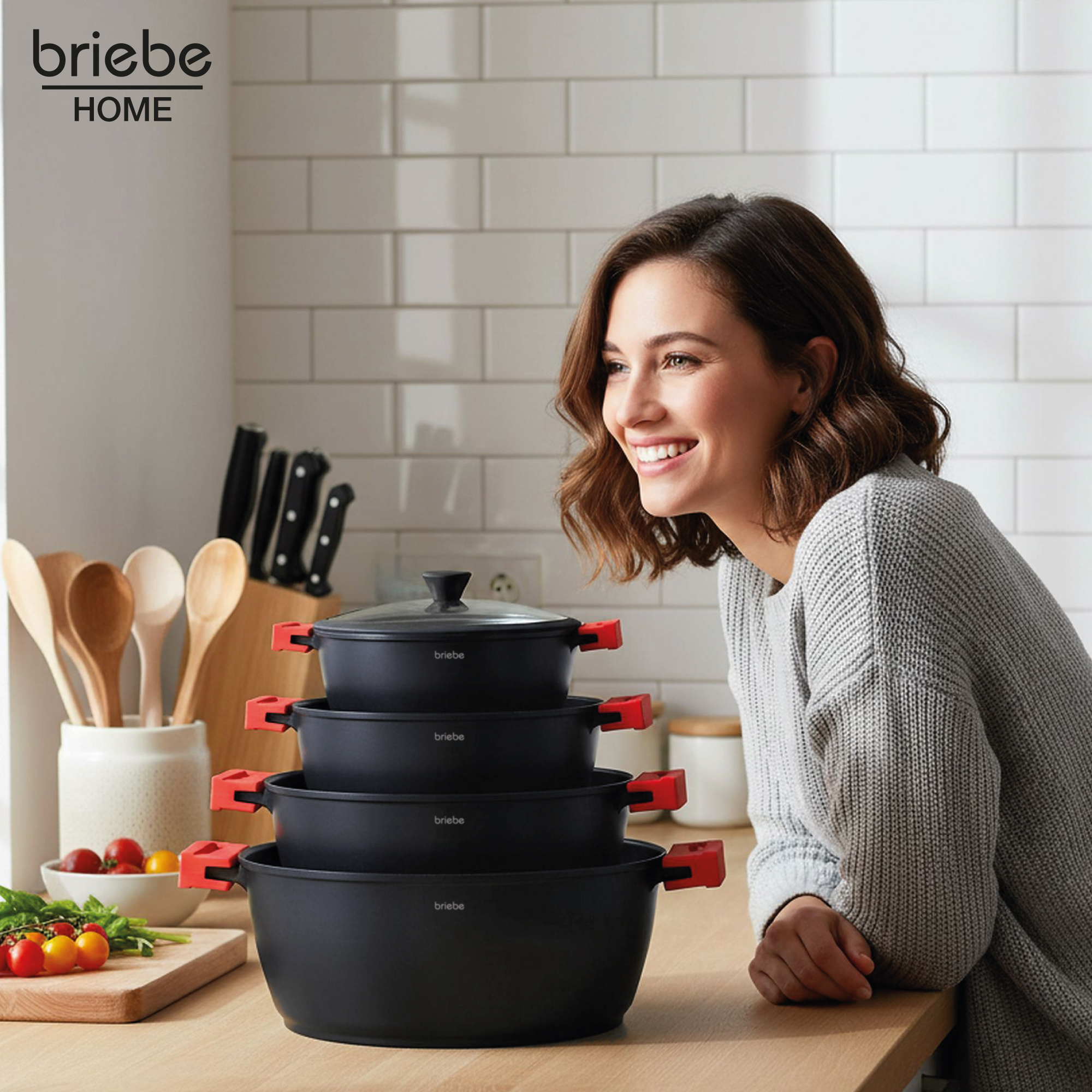 Una mujer sonríe cerca de ollas negras apiladas con asas rojas. Ambiente de cocina con tomates y utensilios de madera.