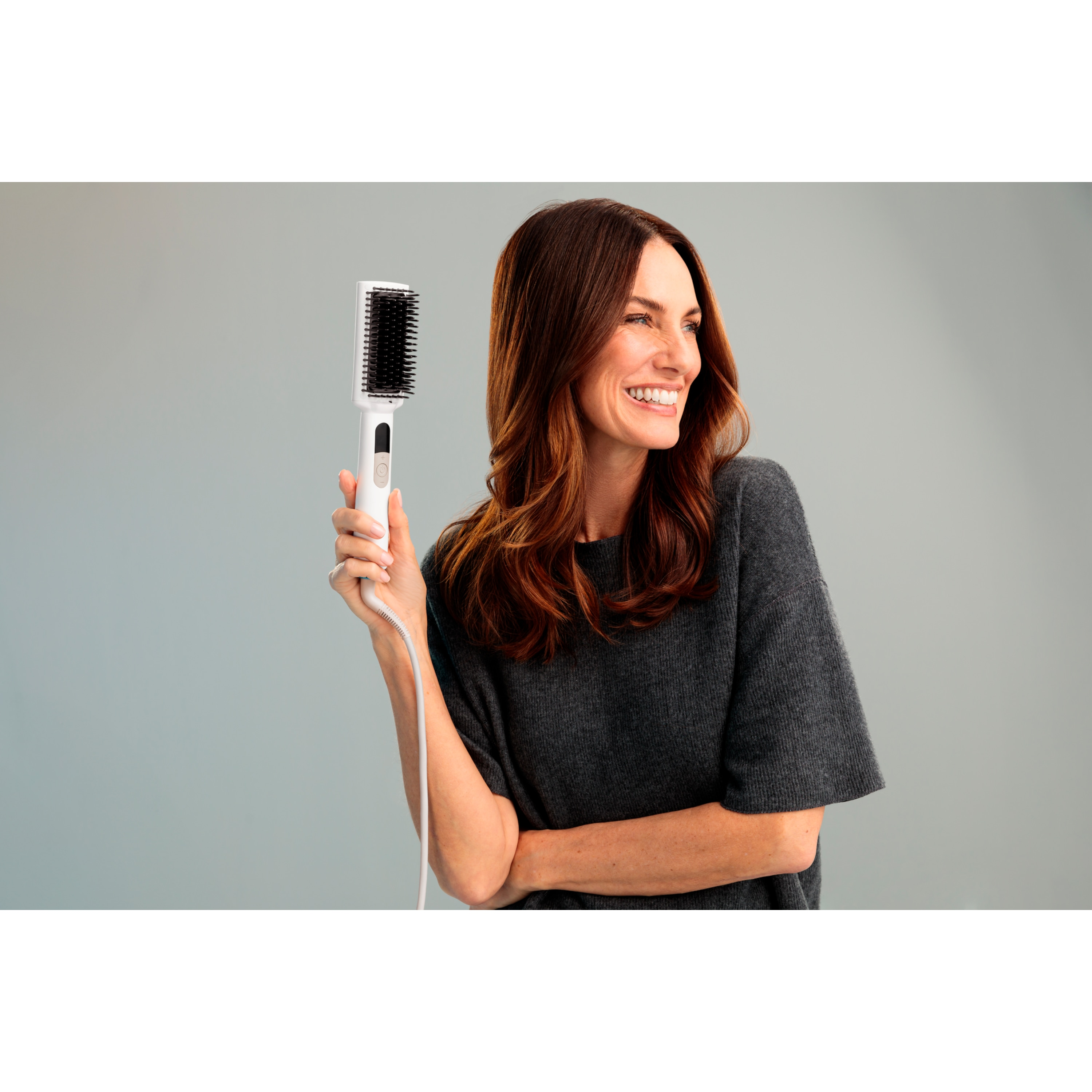 Donna sorridente che tiene una spazzola bianca per capelli. Ha i capelli castani, indossa un maglione grigio e si trova su sfondo grigio.