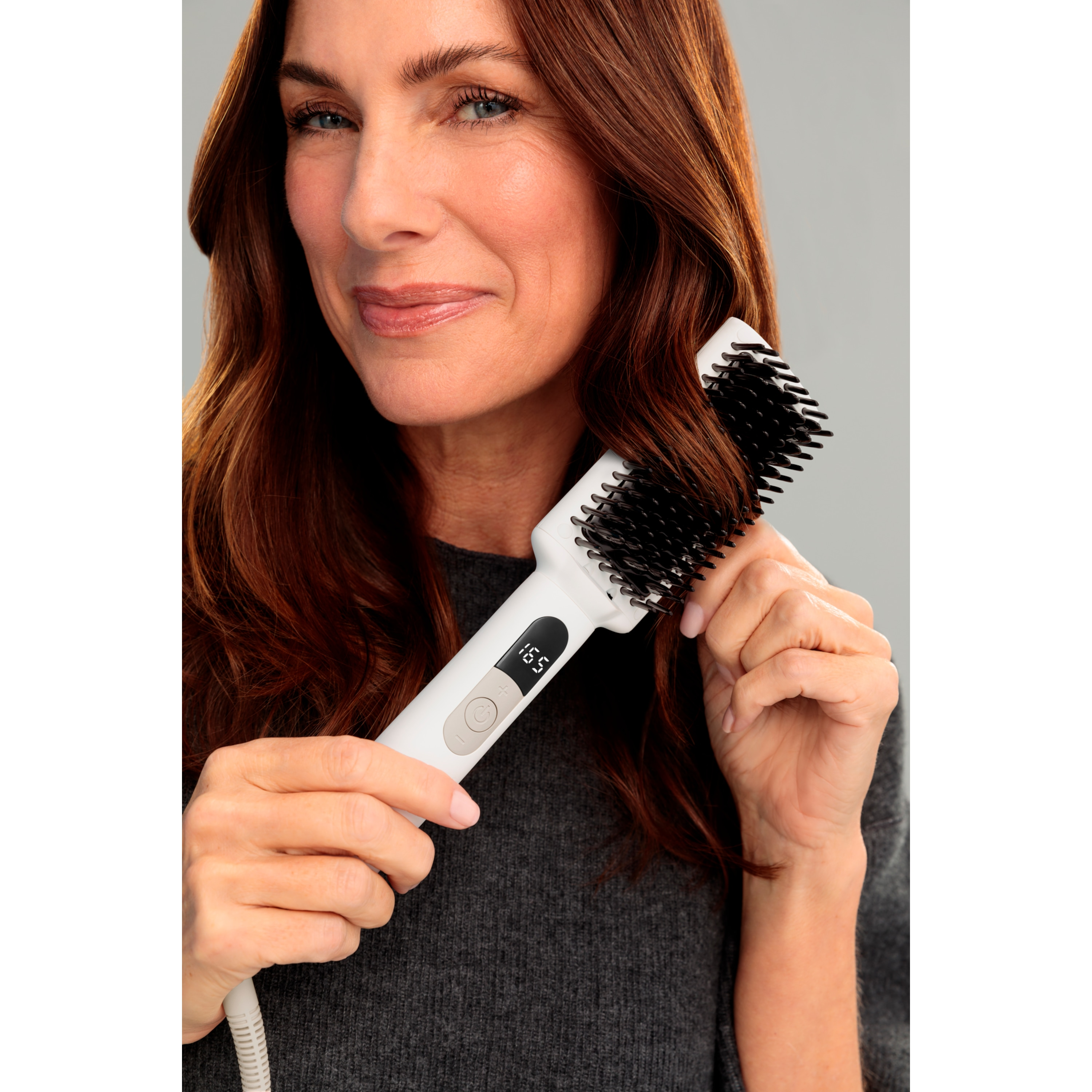 Una donna usa una spazzola bianca per capelli. Ha lunghi capelli castani e indossa un maglione grigio, sorridendo.