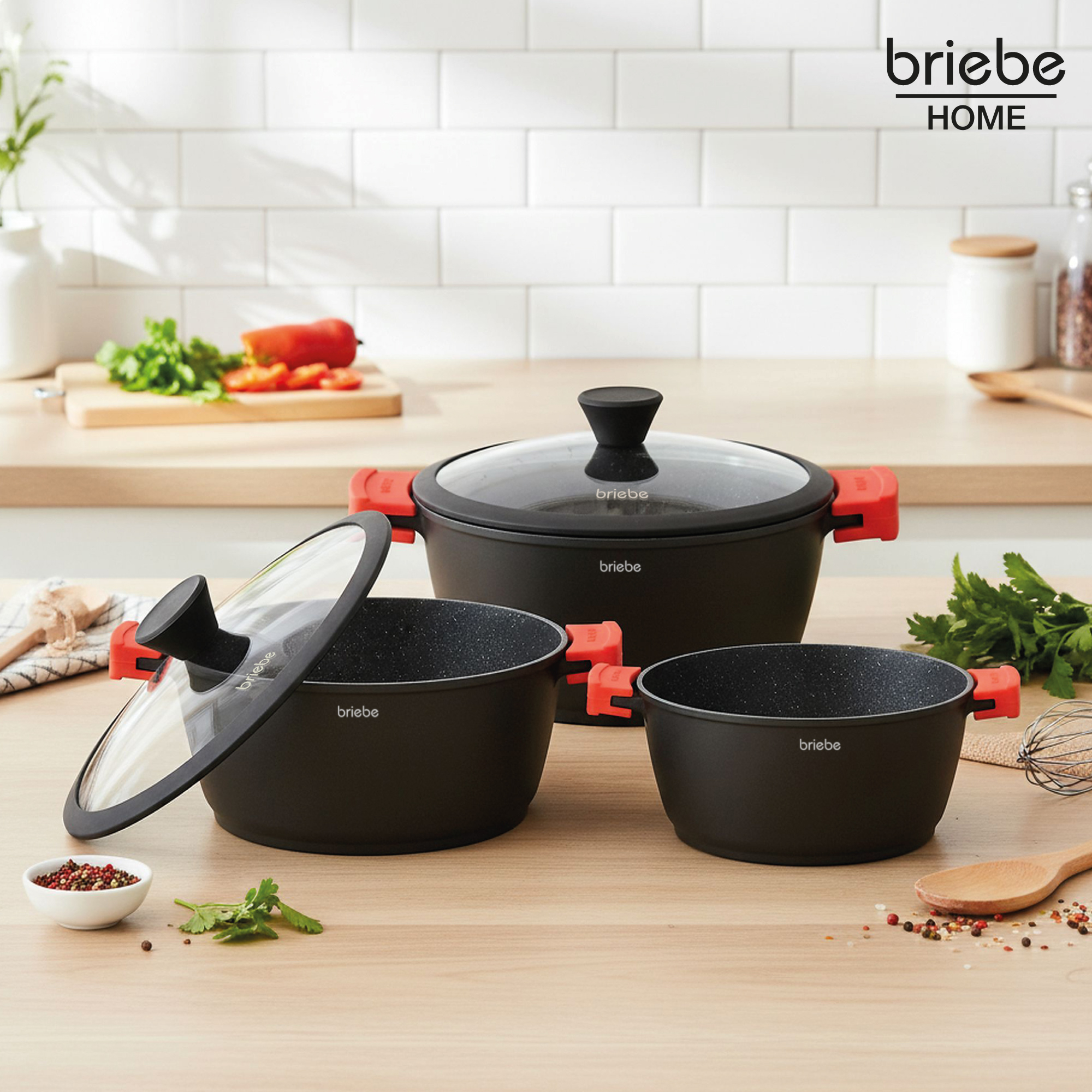 Tres ollas negras con asas rojas, una con tapa. Ambiente de cocina con ingredientes y el logo de Briebe Home.