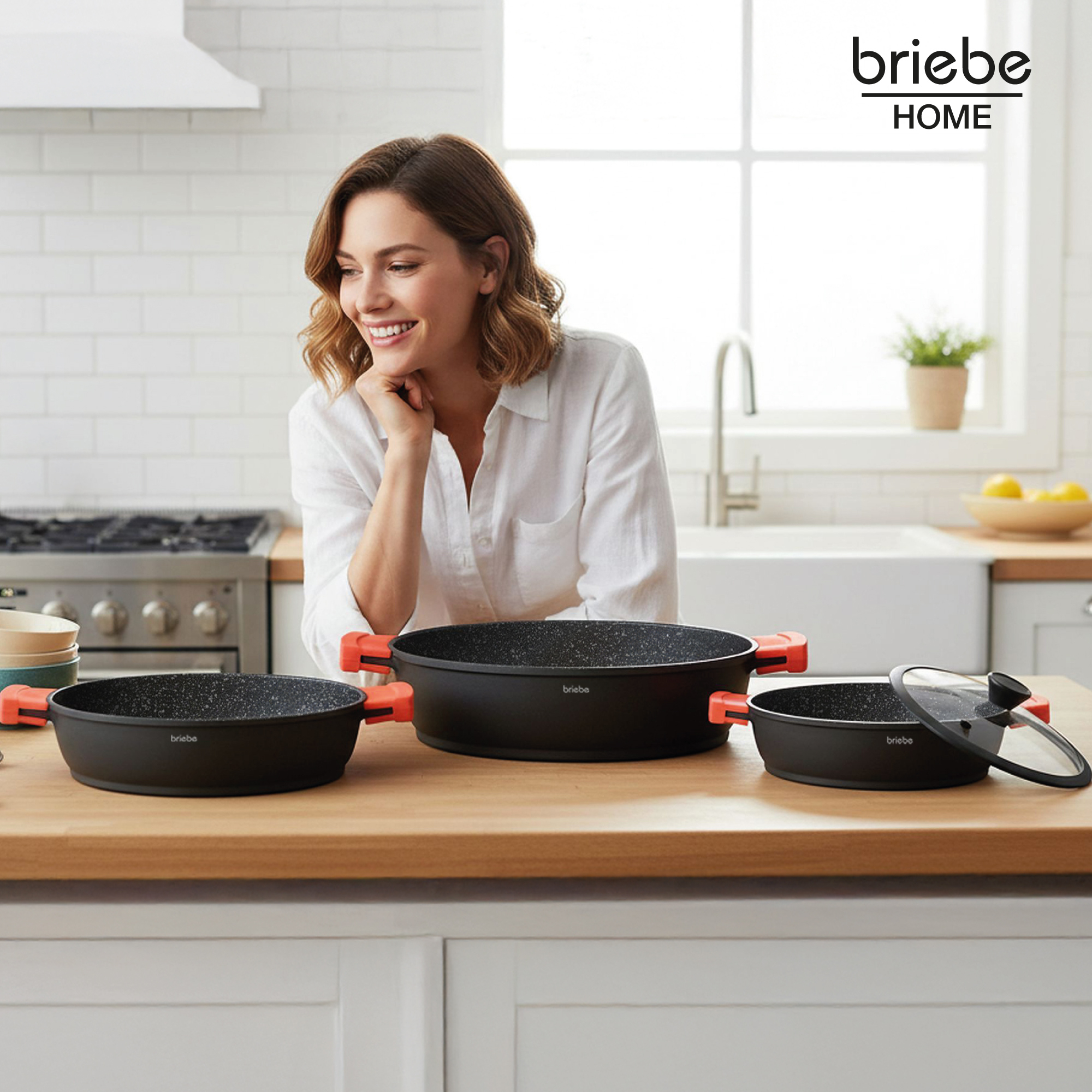 Mujer en cocina con ollas. Sartenes negras con asas naranjas en mostrador de madera. Cocina blanca con logo.