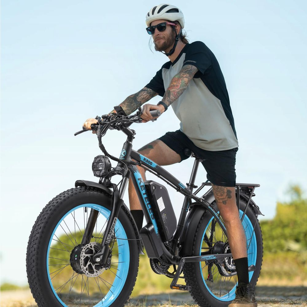 Hombre en una e-bike negra con llantas azules, con casco y gafas de sol. La bicicleta tiene un faro y está en un camino.