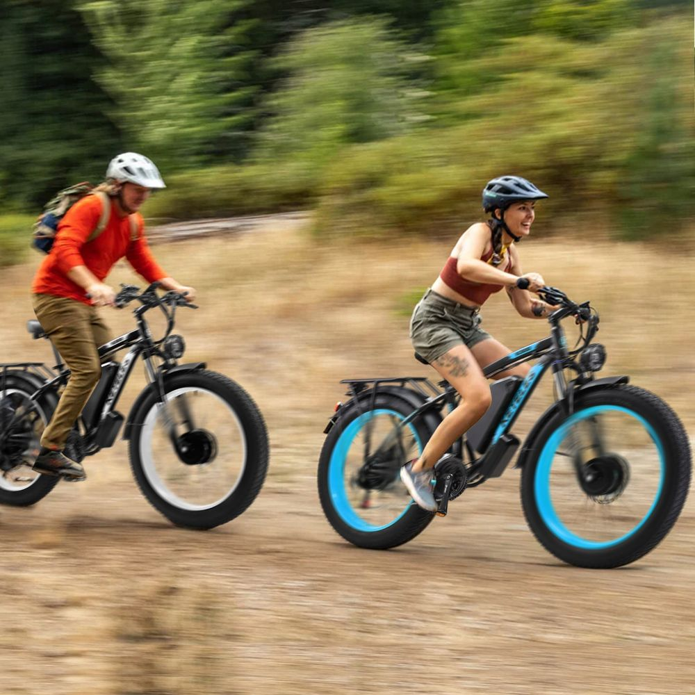 Dos personas montan bicicletas eléctricas de llantas anchas en un camino de tierra. Uno de rojo, otro de marrón. Bosque al fondo.
